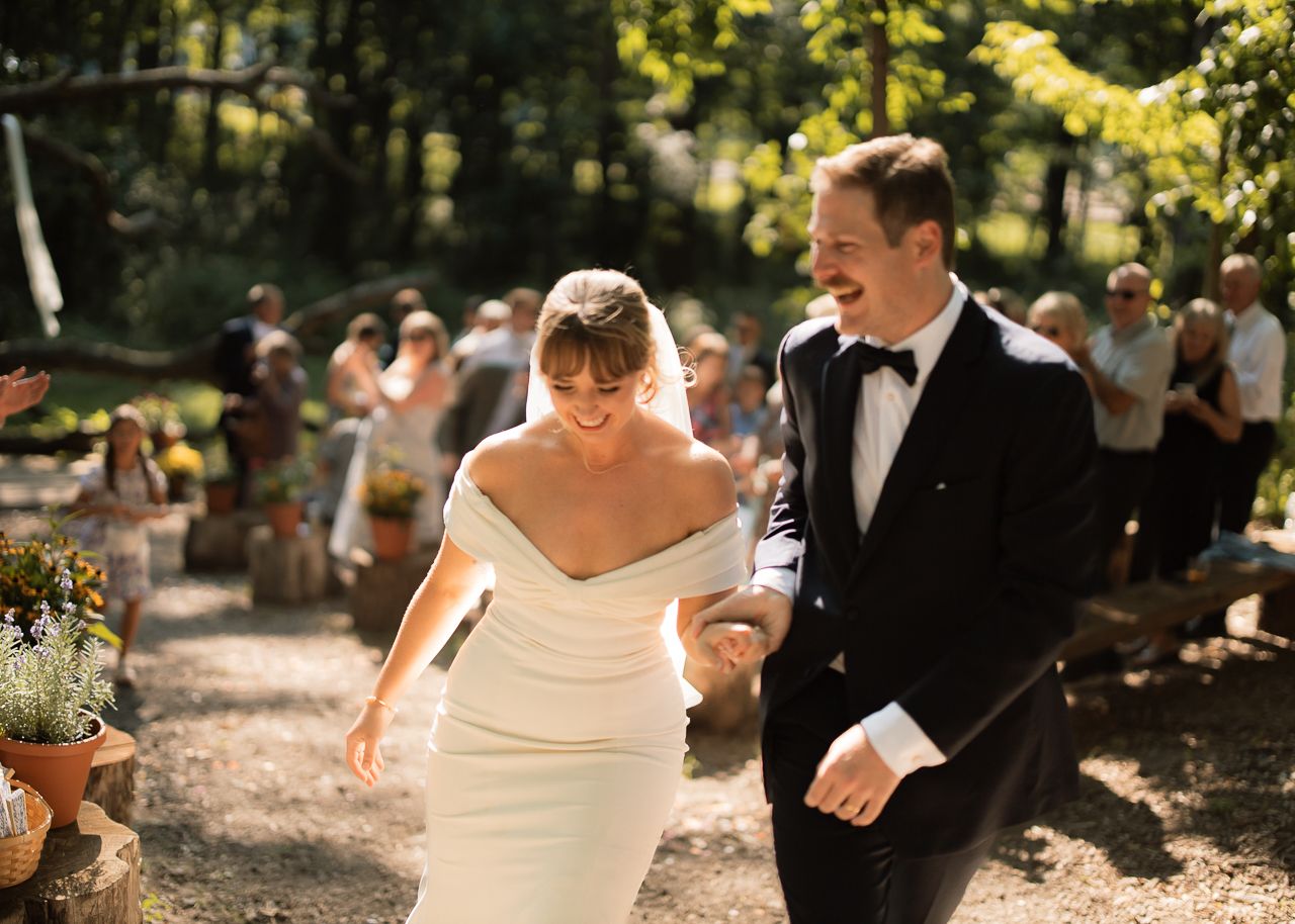 Joyful bride and groom walking down outdoor wedding aisle in elegant white gown and black tuxedo.