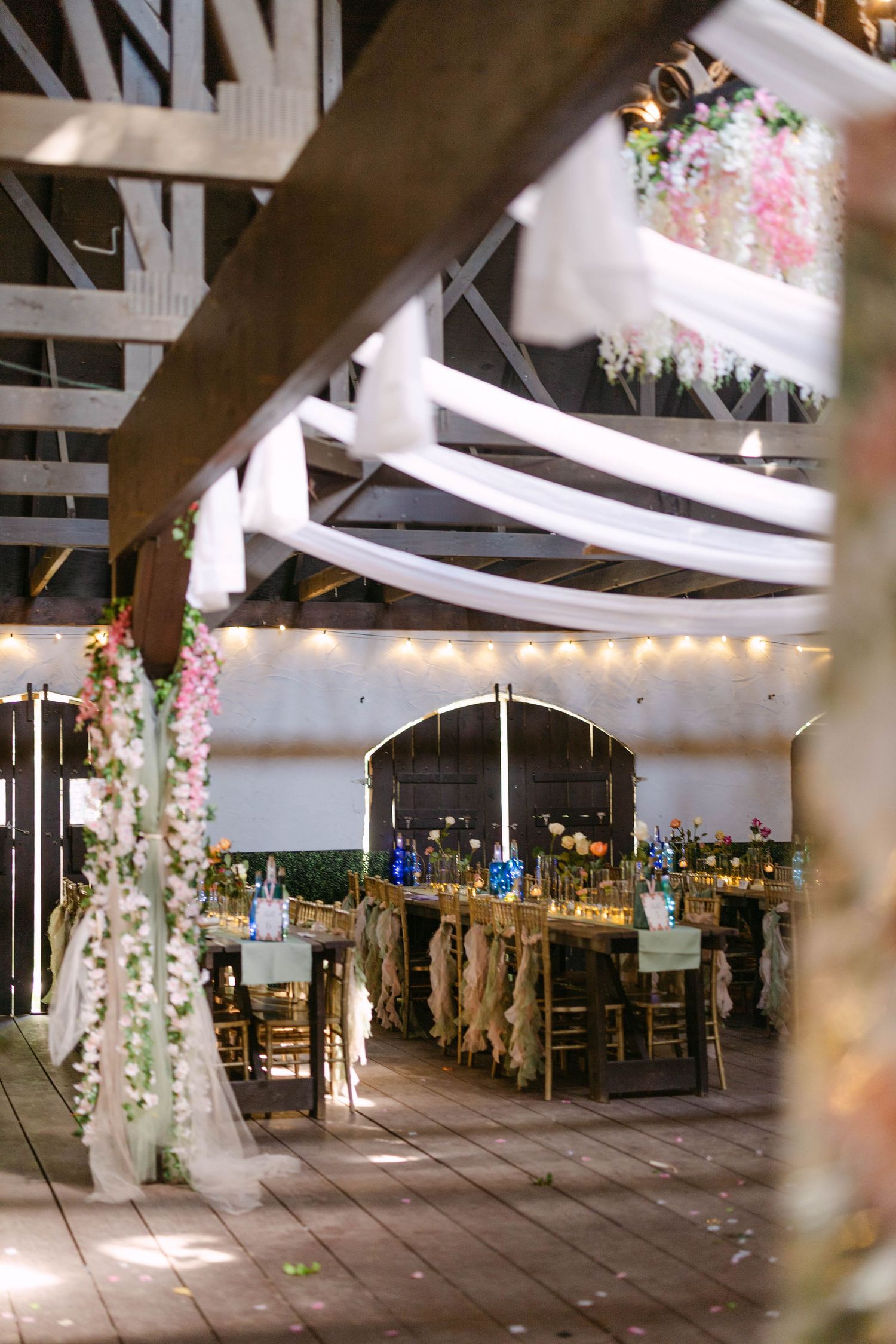 Rustic barn wedding reception with white fabric draping, floral columns, wooden floors, and decorated long dining tables.