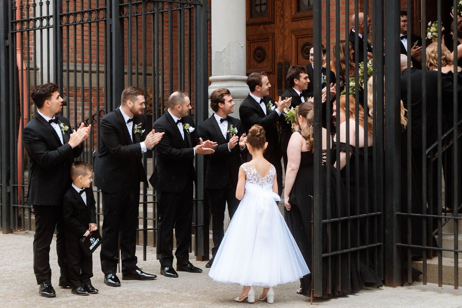 Flower girl in white dress greeted by wedding party in black tuxedos outside elegant venue gates.