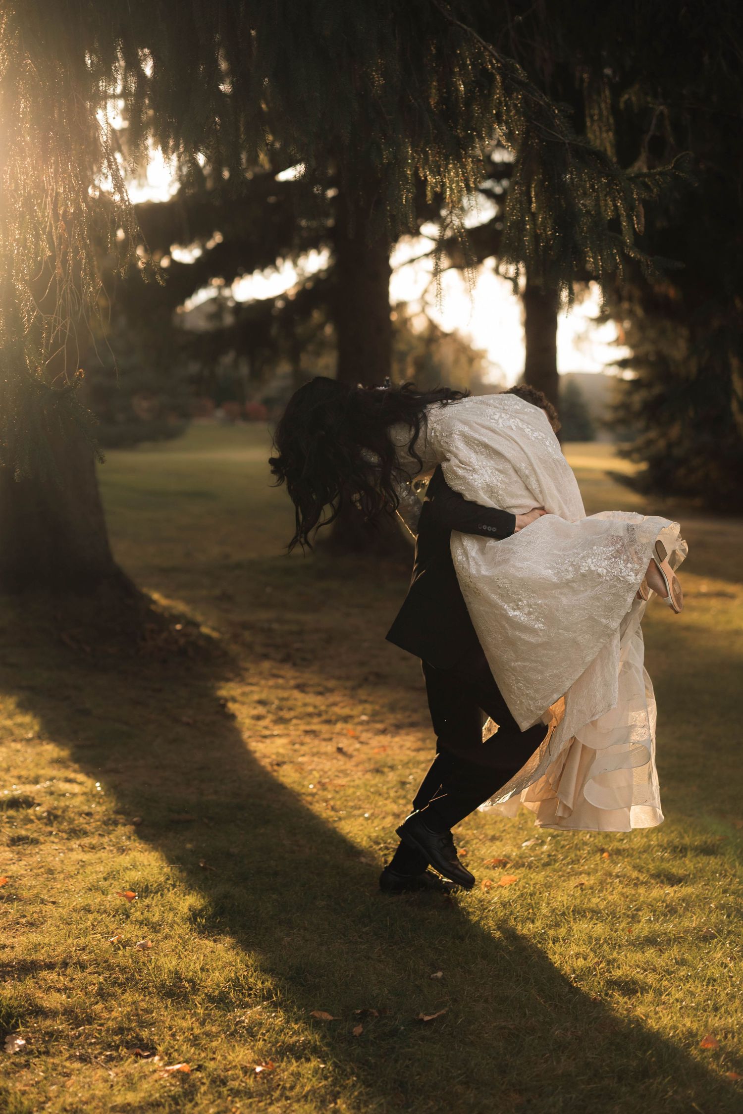 A couple shares a romantic dip kiss in golden sunset light beneath tall trees in a lush green park.