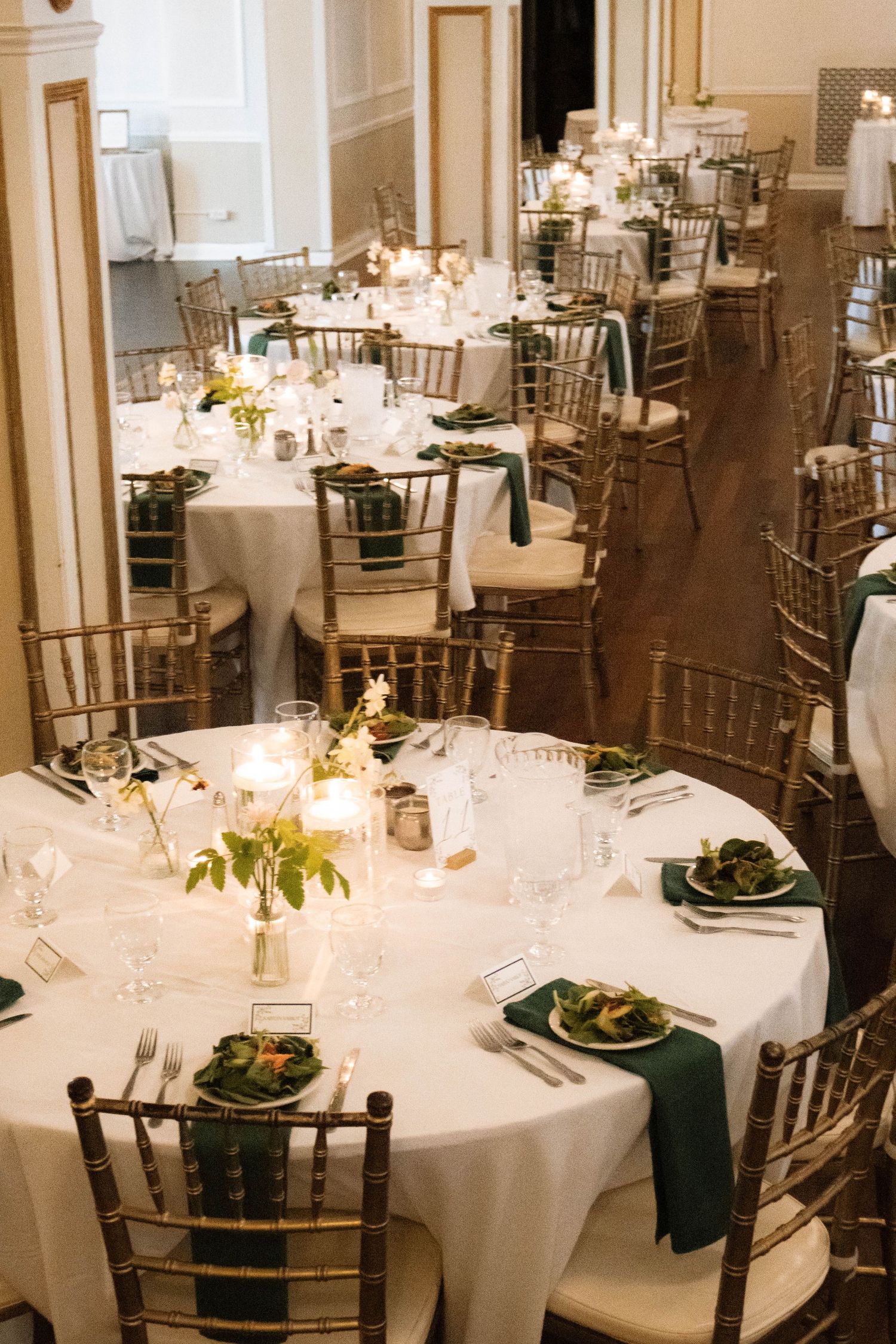 Elegant wedding reception hall with round tables, gold chiavari chairs, and green and white decor.