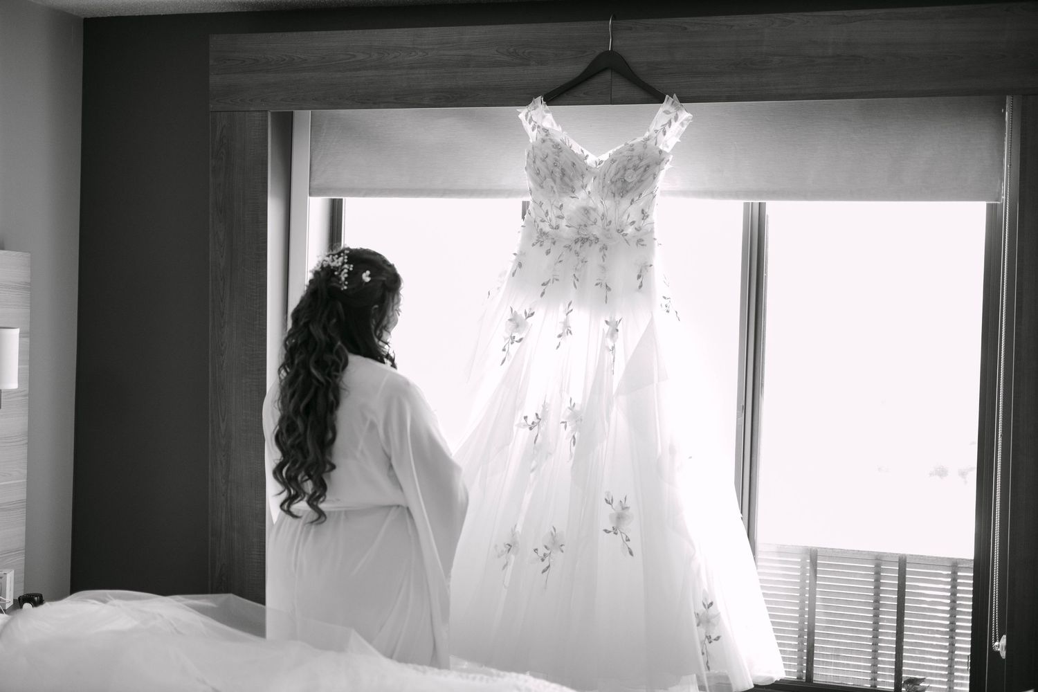 Bride in white robe admiring her elegant lace wedding gown hanging in a hotel room, black and white photo.