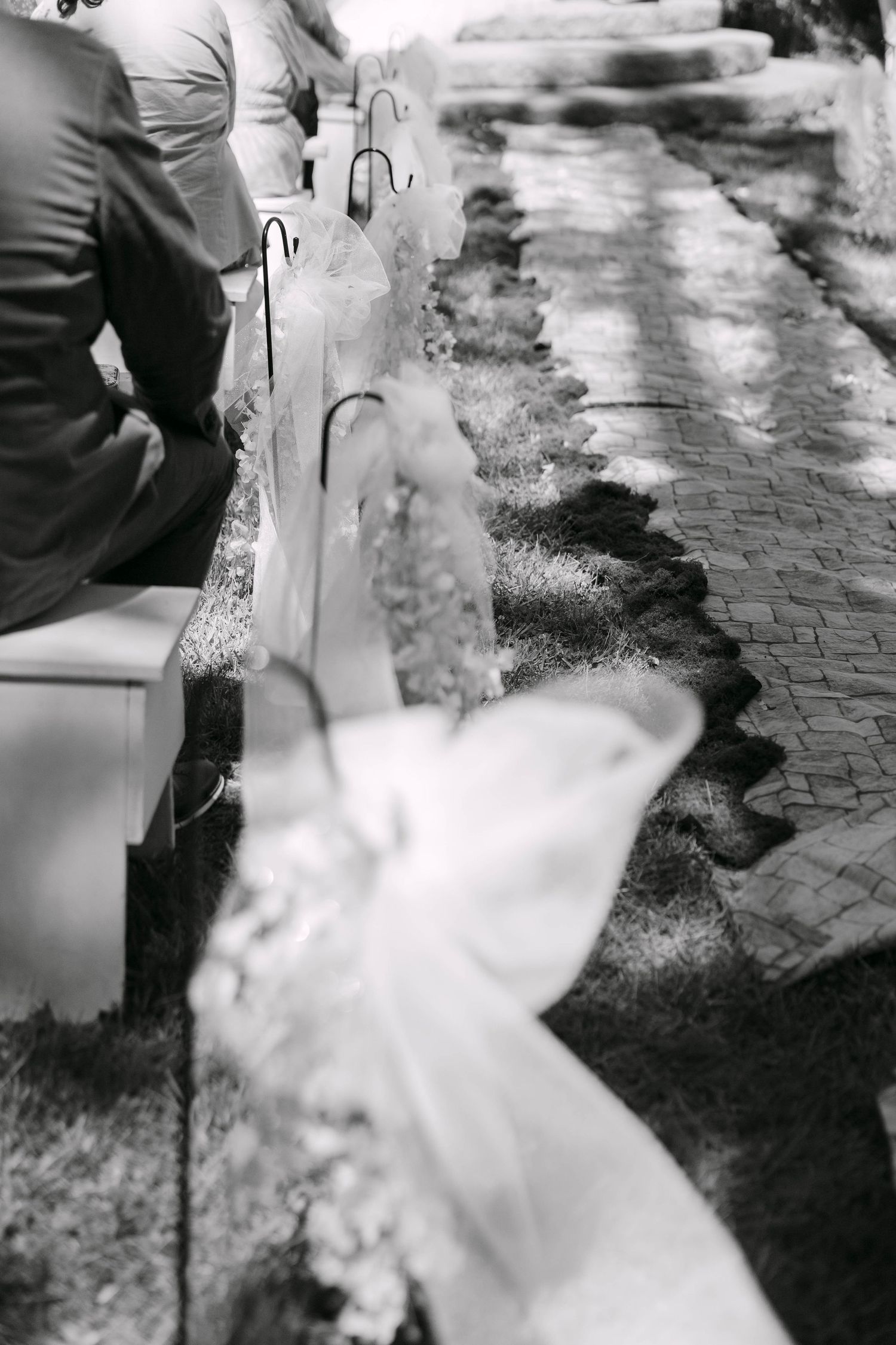 Black and white photo of wedding ceremony aisle decorated with white ribbon bows on chairs along a brick pathway.