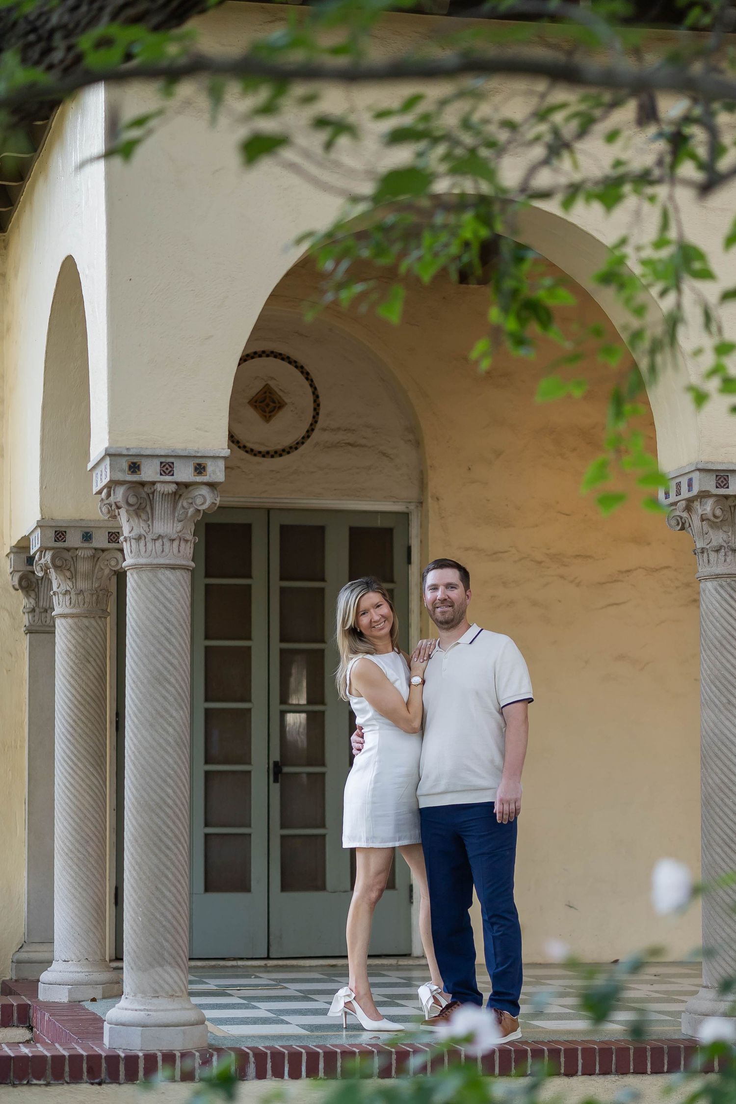 Couple posing under ornate arched colonnade of Mediterranean-style building in white dress and polo shirt.