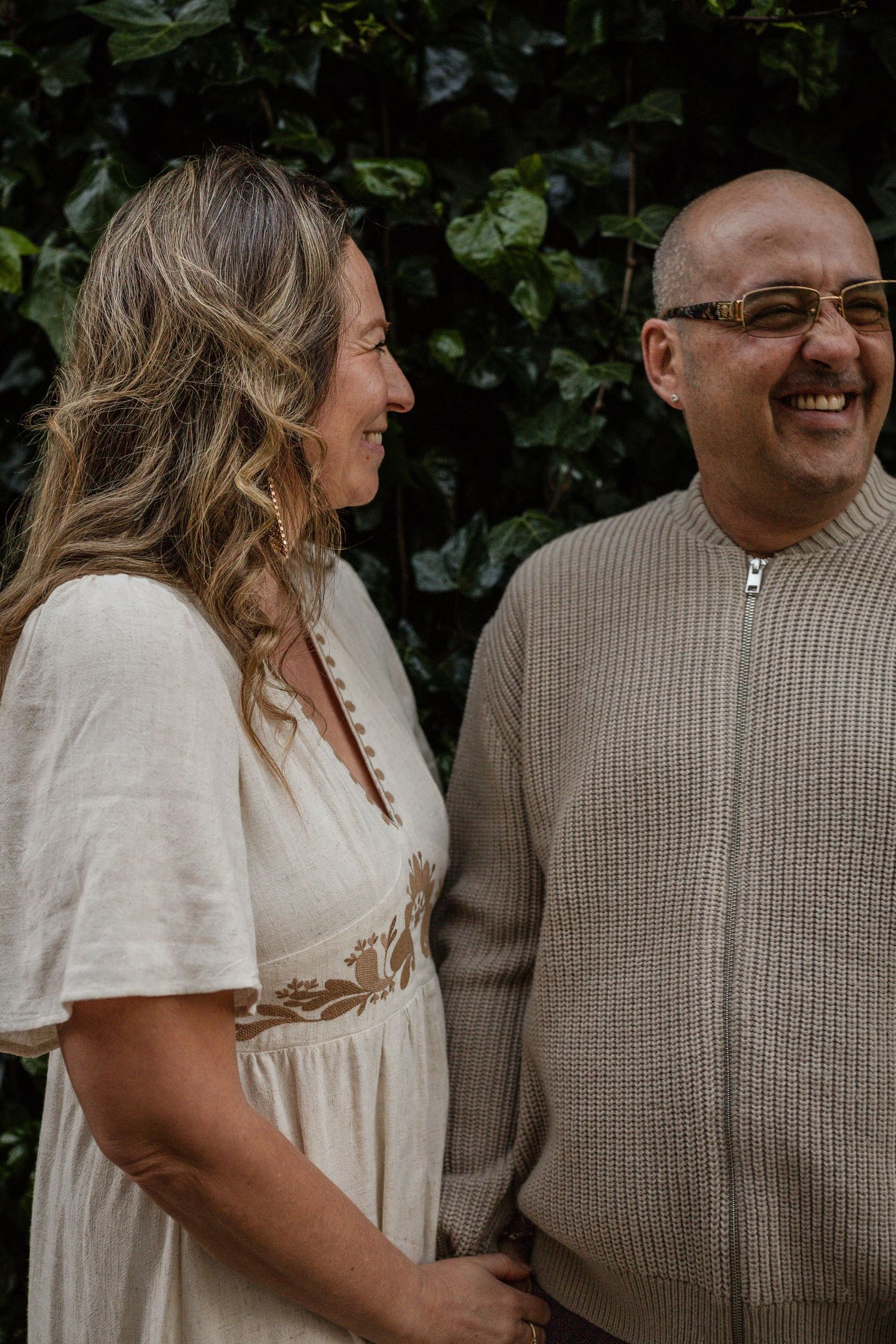 A smiling bald man with glasses and a woman in a white dress laugh together outdoors near green foliage.