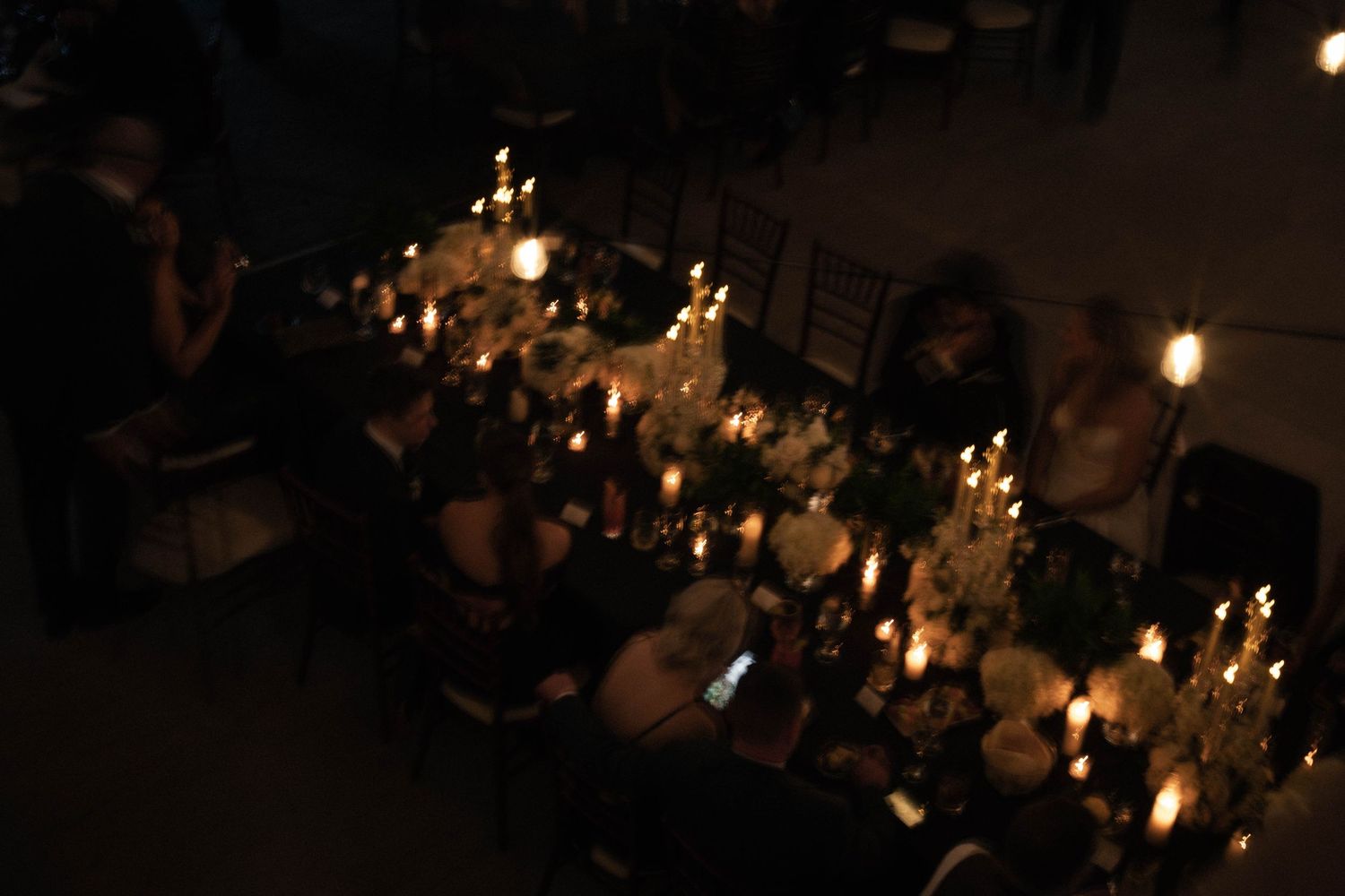 Elegant candlelit dinner party with guests seated at a long decorated table in a dimly lit venue.