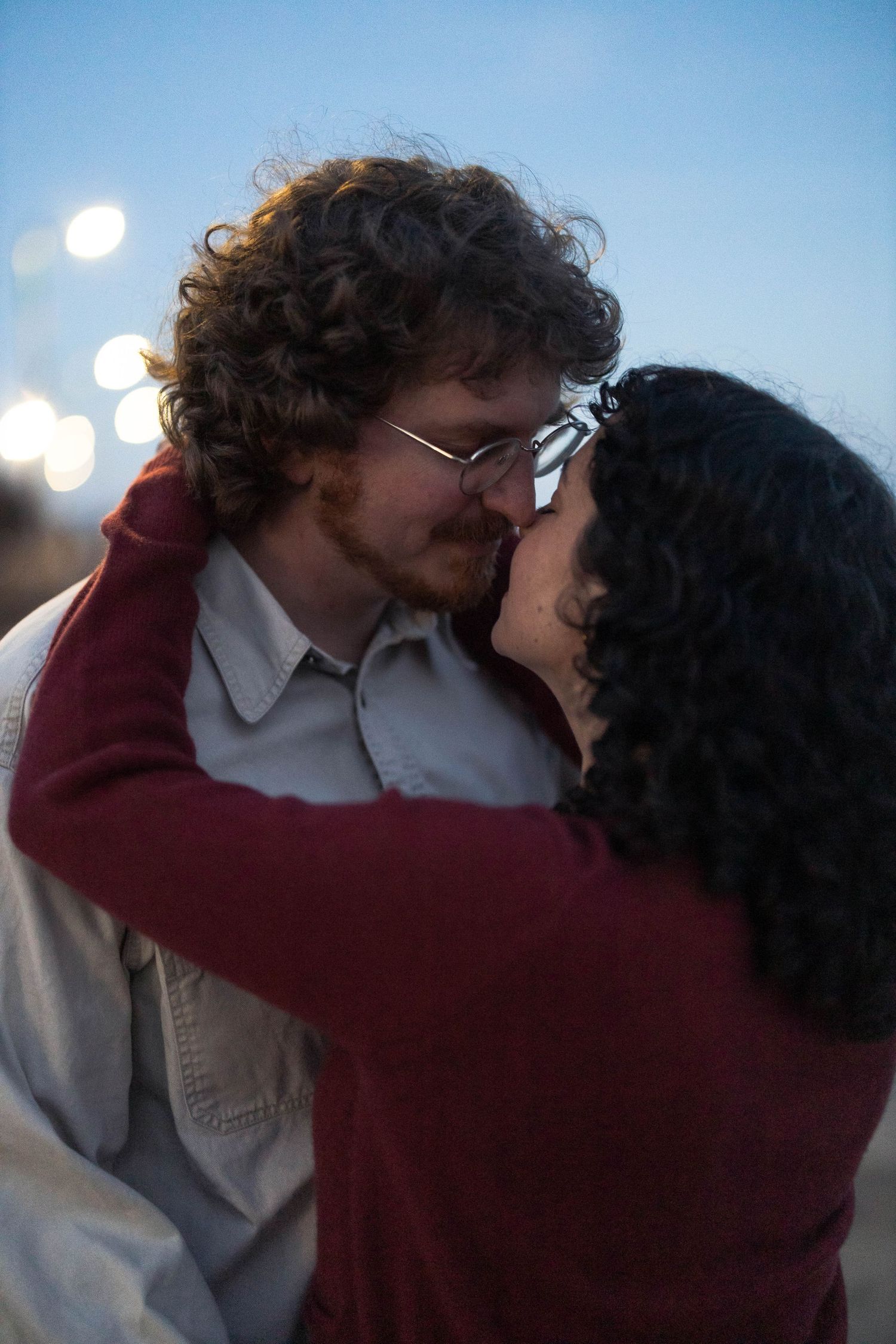 A couple sharing a romantic almost-kiss moment outdoors at dusk, man with curly hair and glasses, woman in red sweater.
