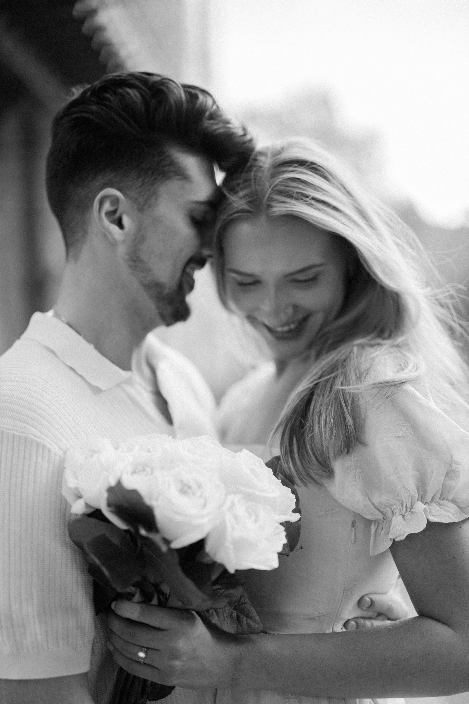 Black and white photo of a smiling couple sharing a romantic moment while holding a bouquet of white roses.