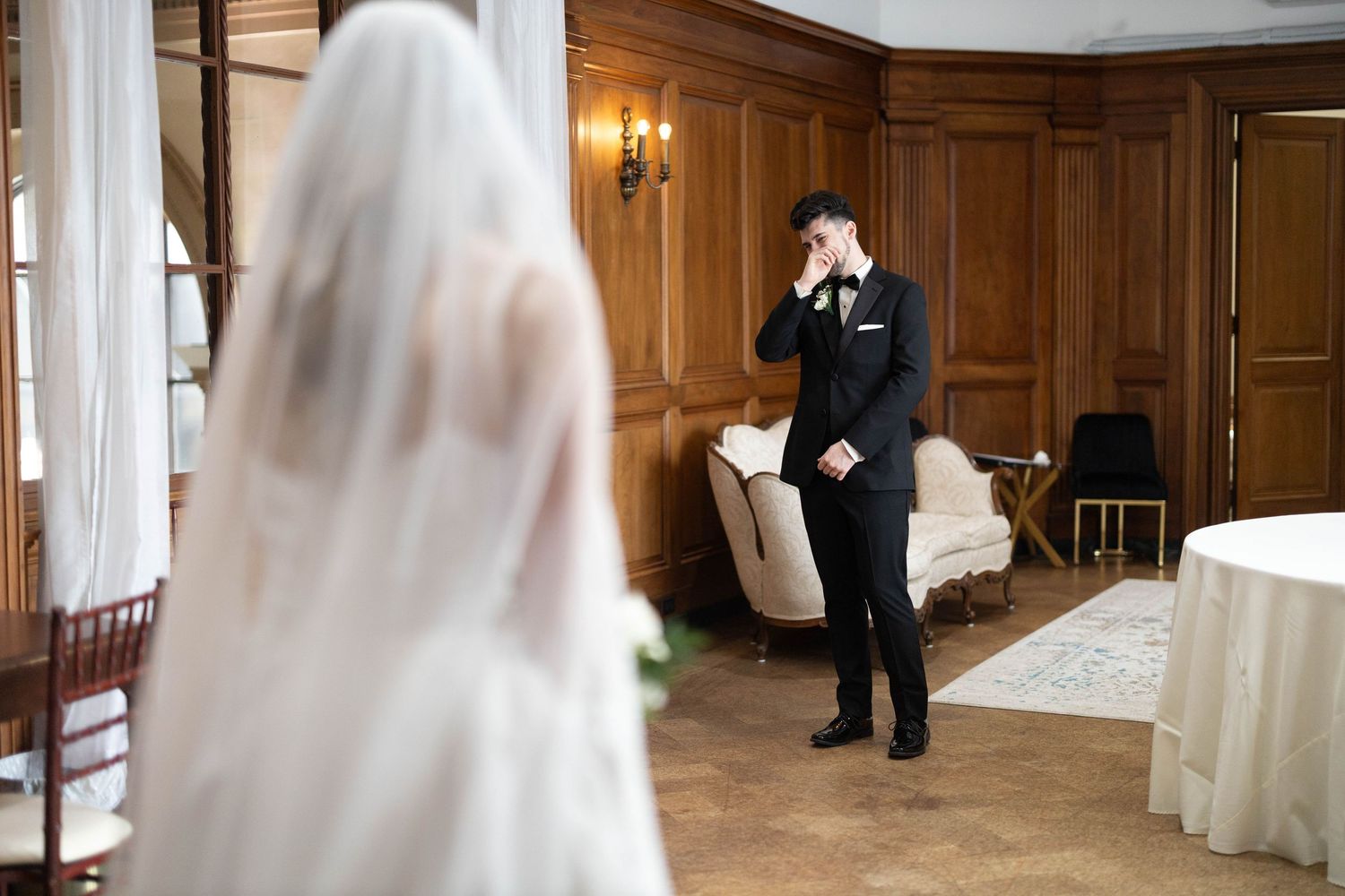 Emotional groom wiping tears during wedding first look moment in elegant wood-paneled room with bride in white veil.