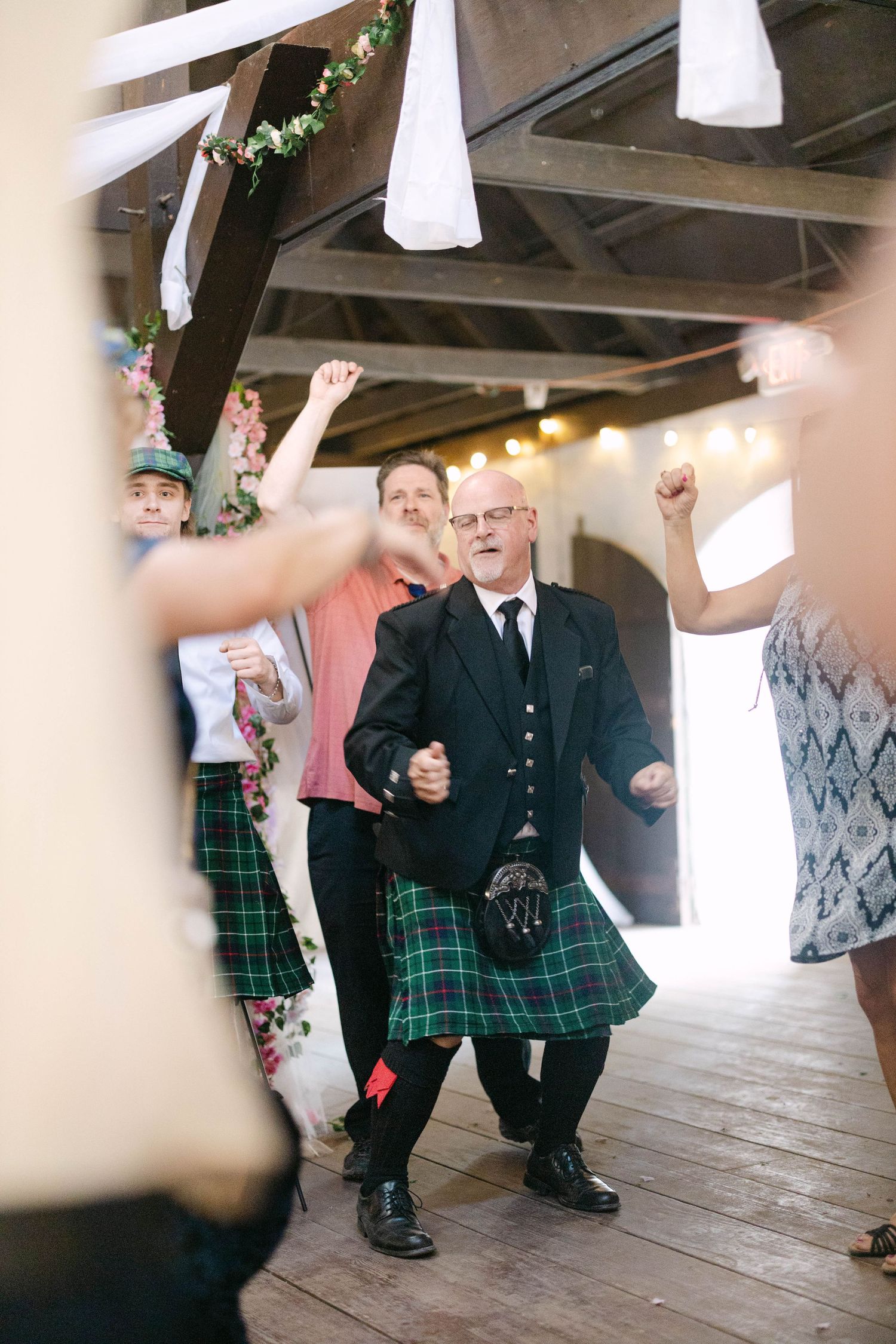 Man in traditional Scottish kilt dancing joyfully at a wedding reception in a rustic barn venue.