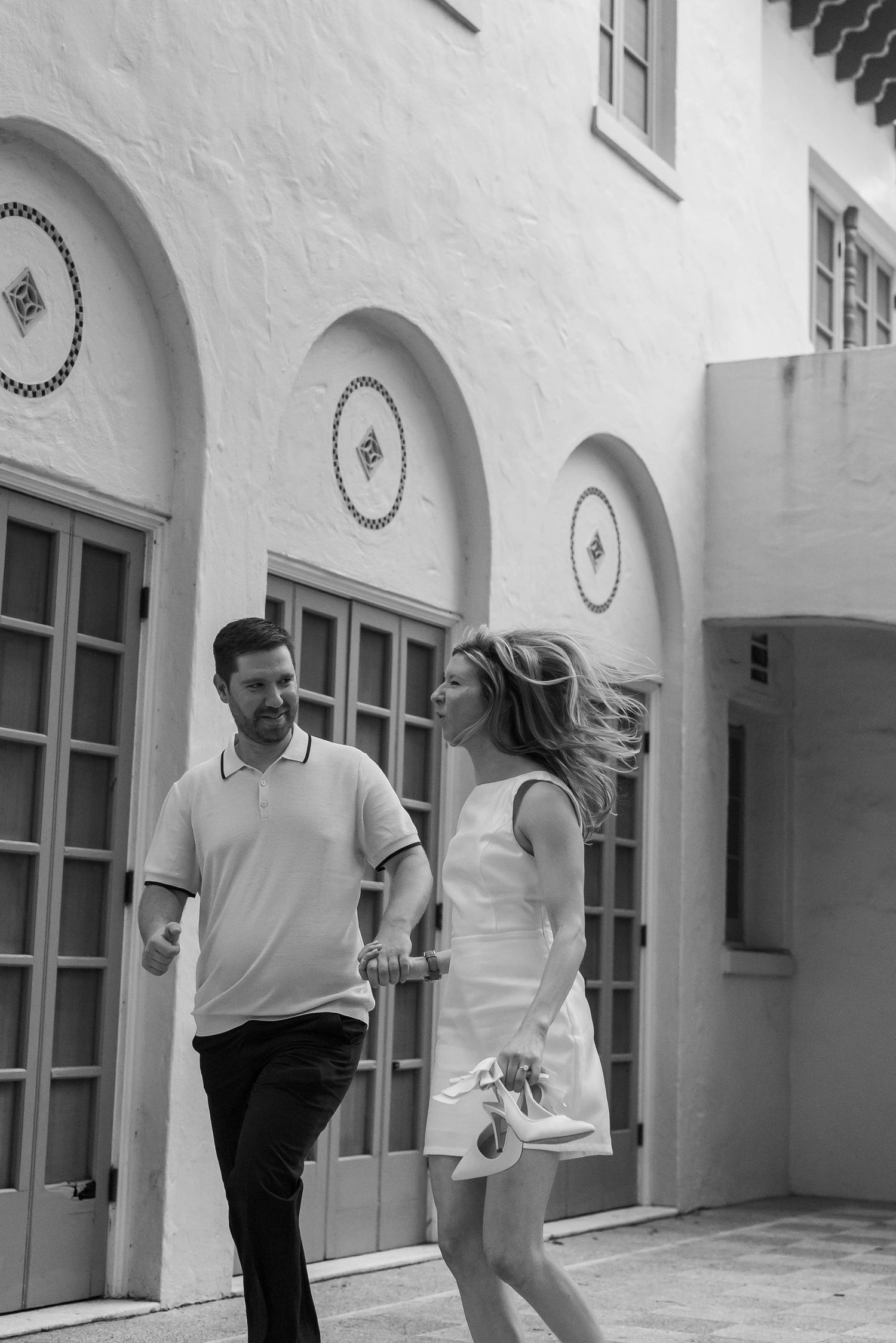 Black and white photo of couple walking and laughing outside Mediterranean architecture with arched windows.