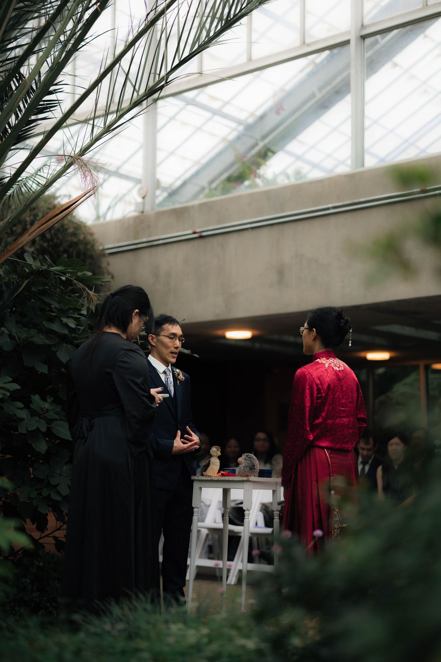 Wedding ceremony in botanical greenhouse with groom, officiant, and bride in red dress.