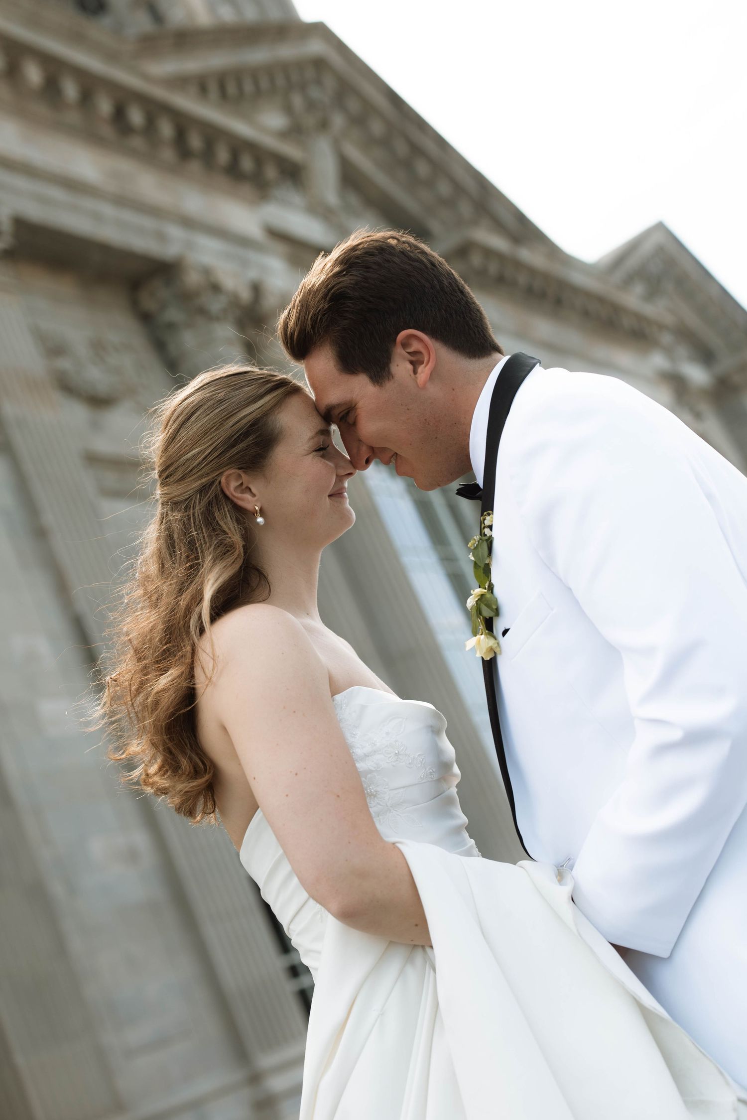 Bride and groom share a romantic forehead touch in wedding attire outside a classic stone building.
