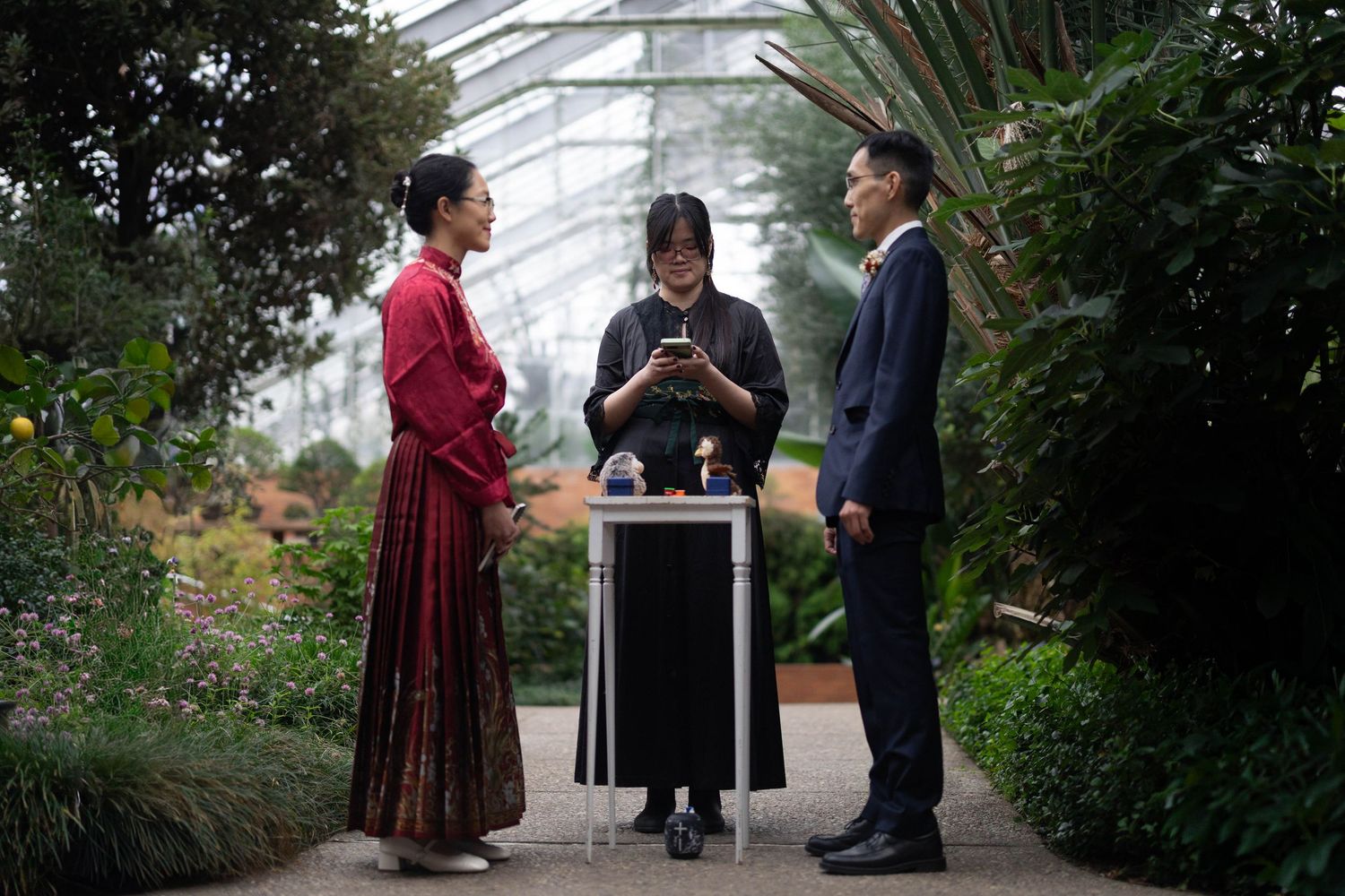 Three people in elegant attire converse in a lush greenhouse, with one person checking a smartphone at a podium.