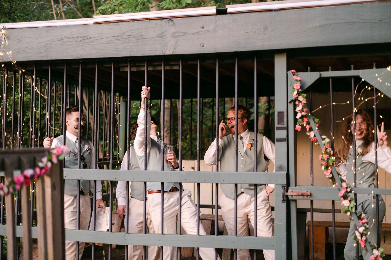 Groomsmen in tan suits laughing behind decorative iron gates adorned with floral garlands.