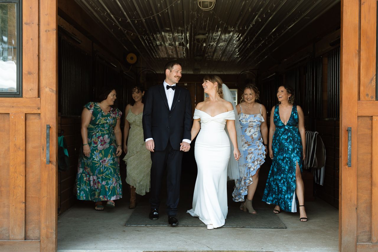 Bride and groom walk hand-in-hand through barn doors with bridesmaids in floral gowns at elegant wedding.
