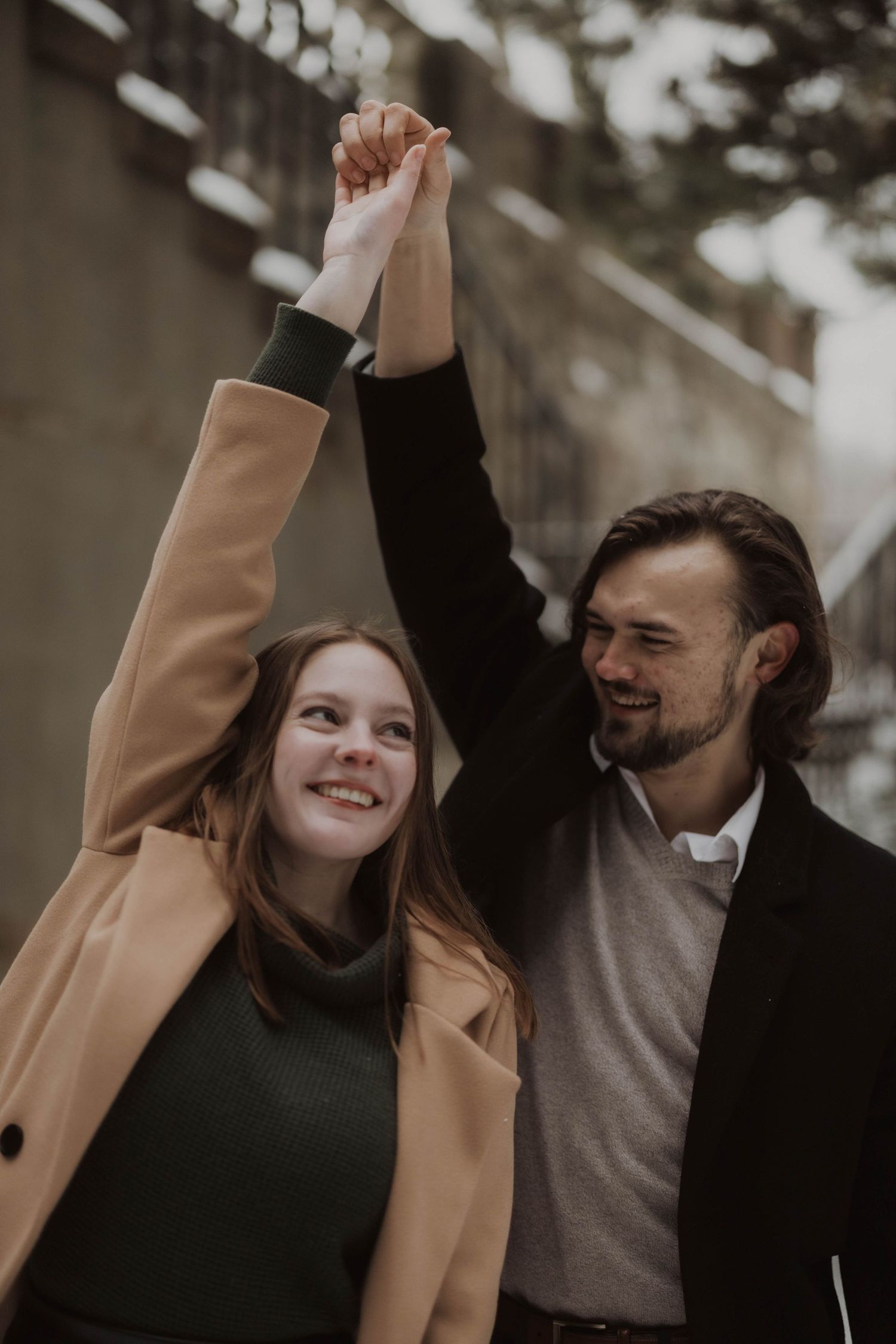 A smiling couple raises their joined hands together outdoors, woman in tan coat and man in black jacket.