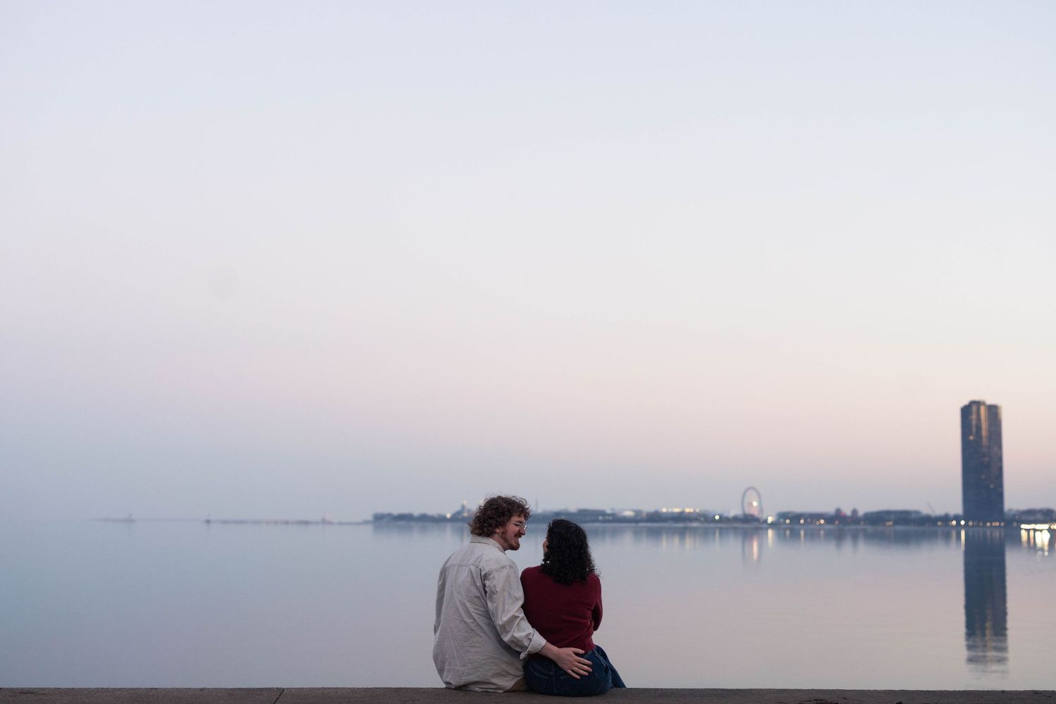 Couple sitting together on a waterfront ledge at dusk, enjoying a serene pastel sky over calm water.