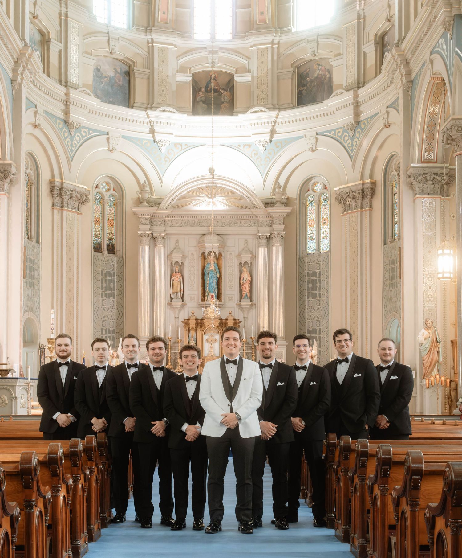 Groom in white tuxedo stands with groomsmen in black tuxedos inside an ornate domed Catholic church.