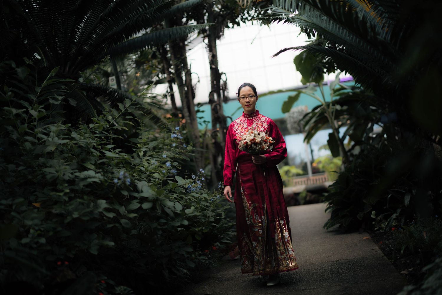 Woman in traditional red floral qipao dress walking along a lush tropical greenhouse garden pathway.