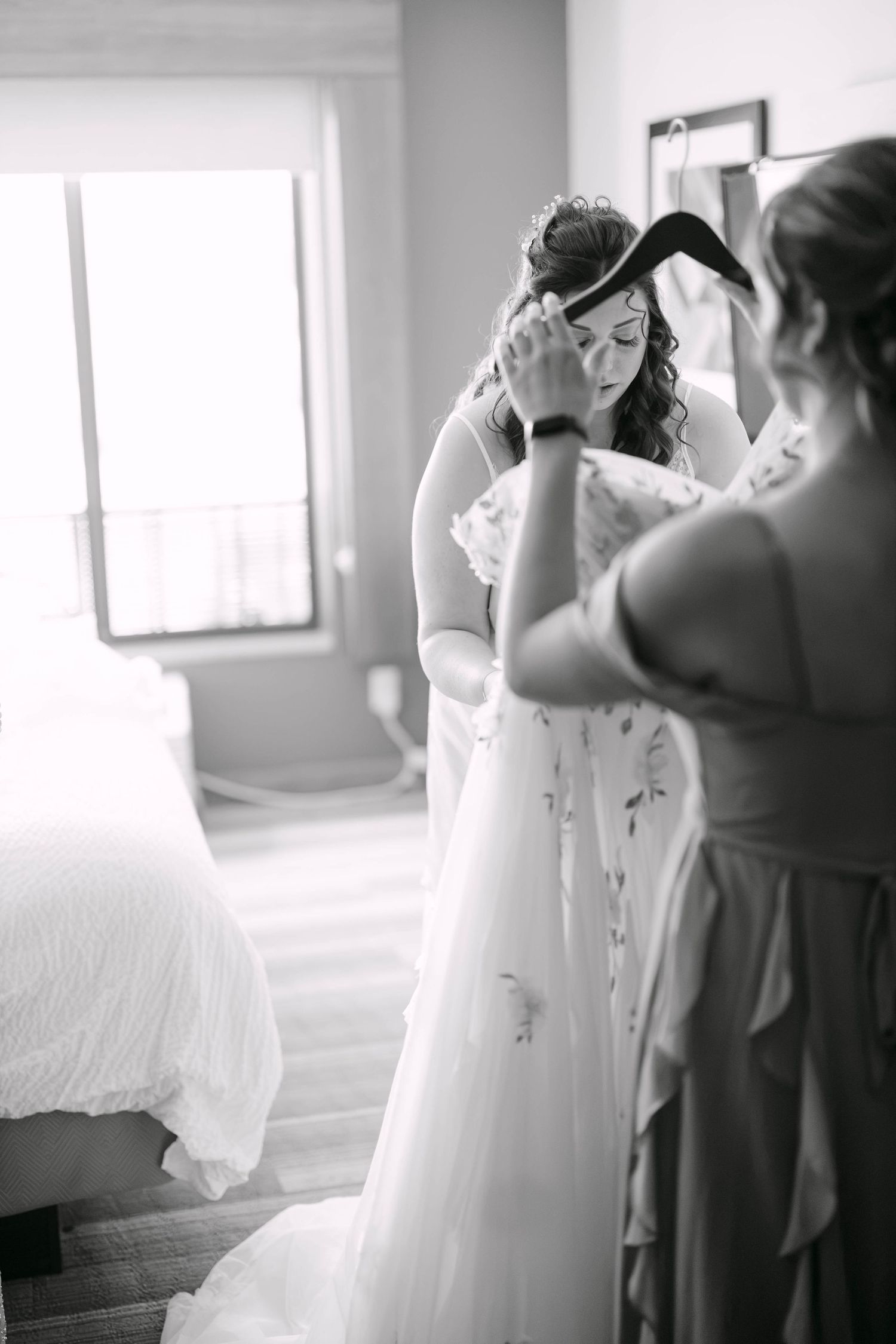 Black and white photo of a bride being helped into her wedding dress by a bridesmaid in a bright hotel room.