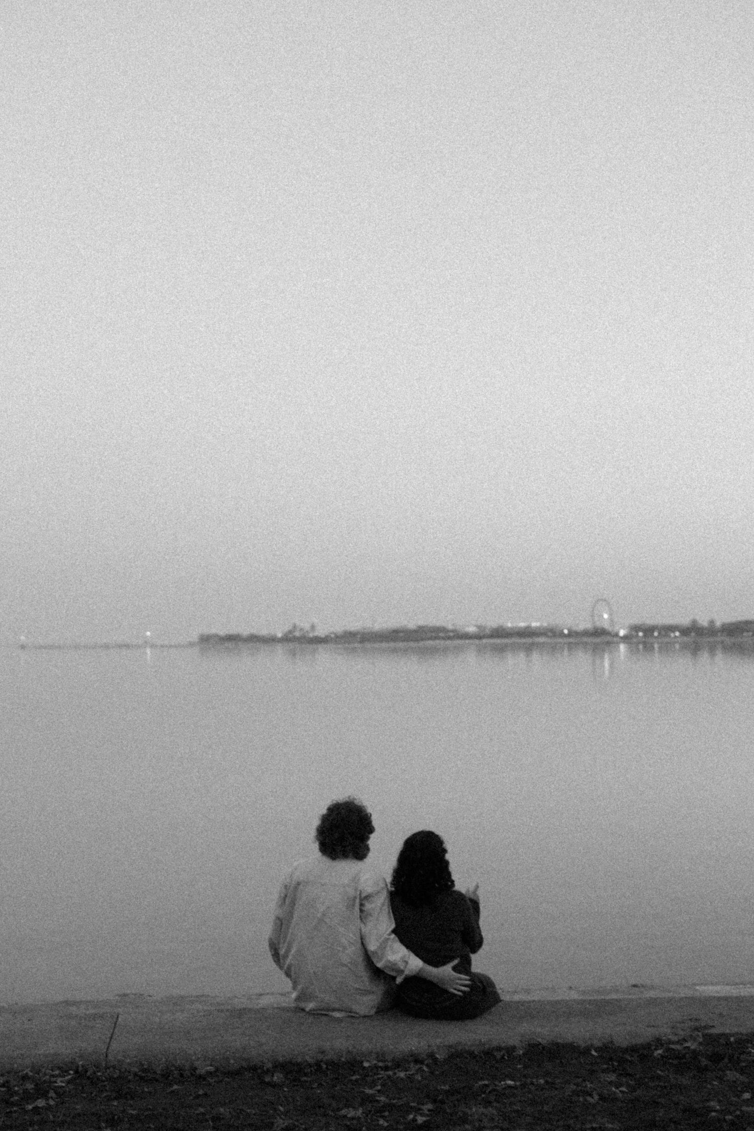 Couple sitting together on waterfront at dusk in black and white romantic photo.