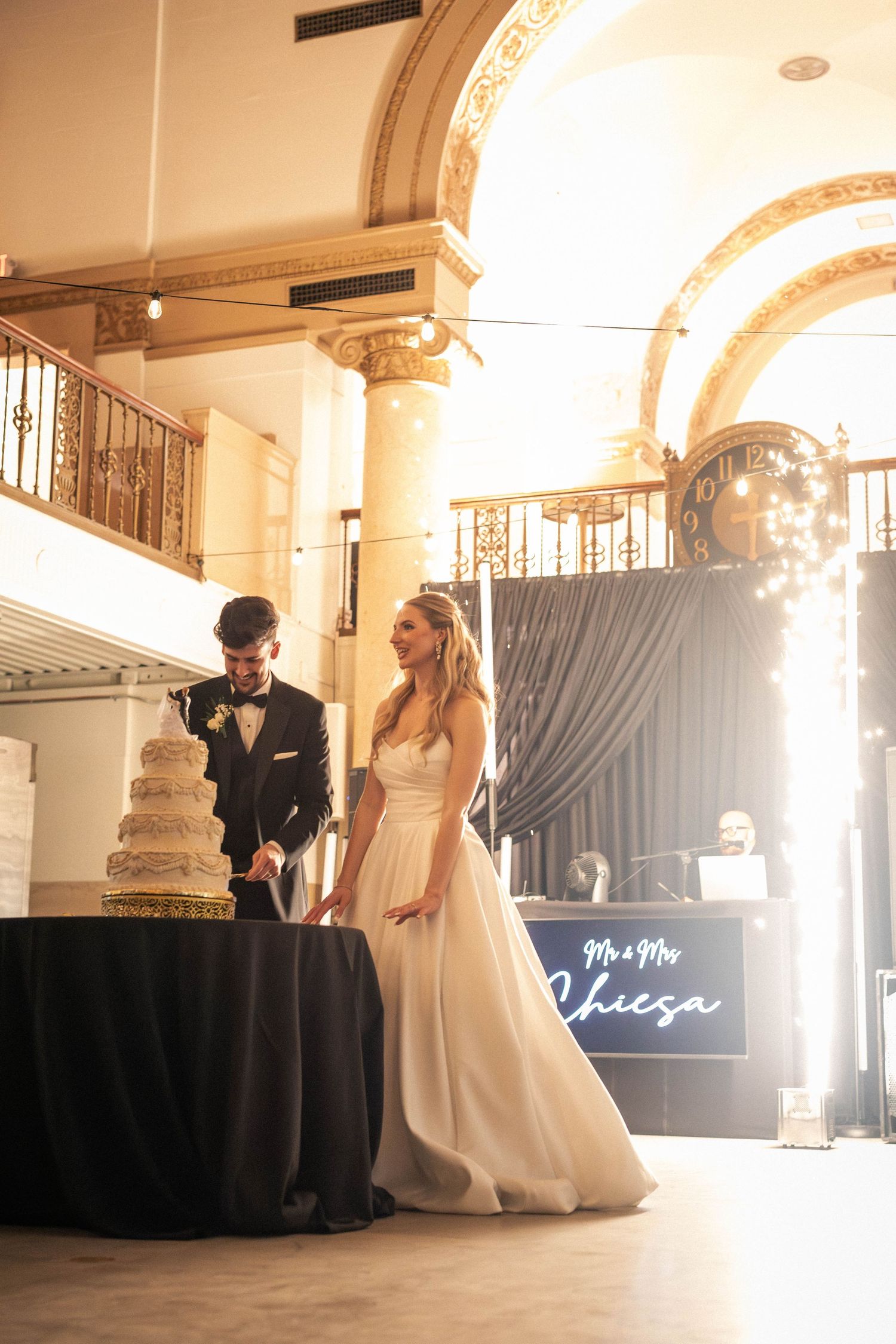 Bride and groom cutting wedding cake in elegant venue with arched columns and Mr & Mrs Chiesa neon sign.
