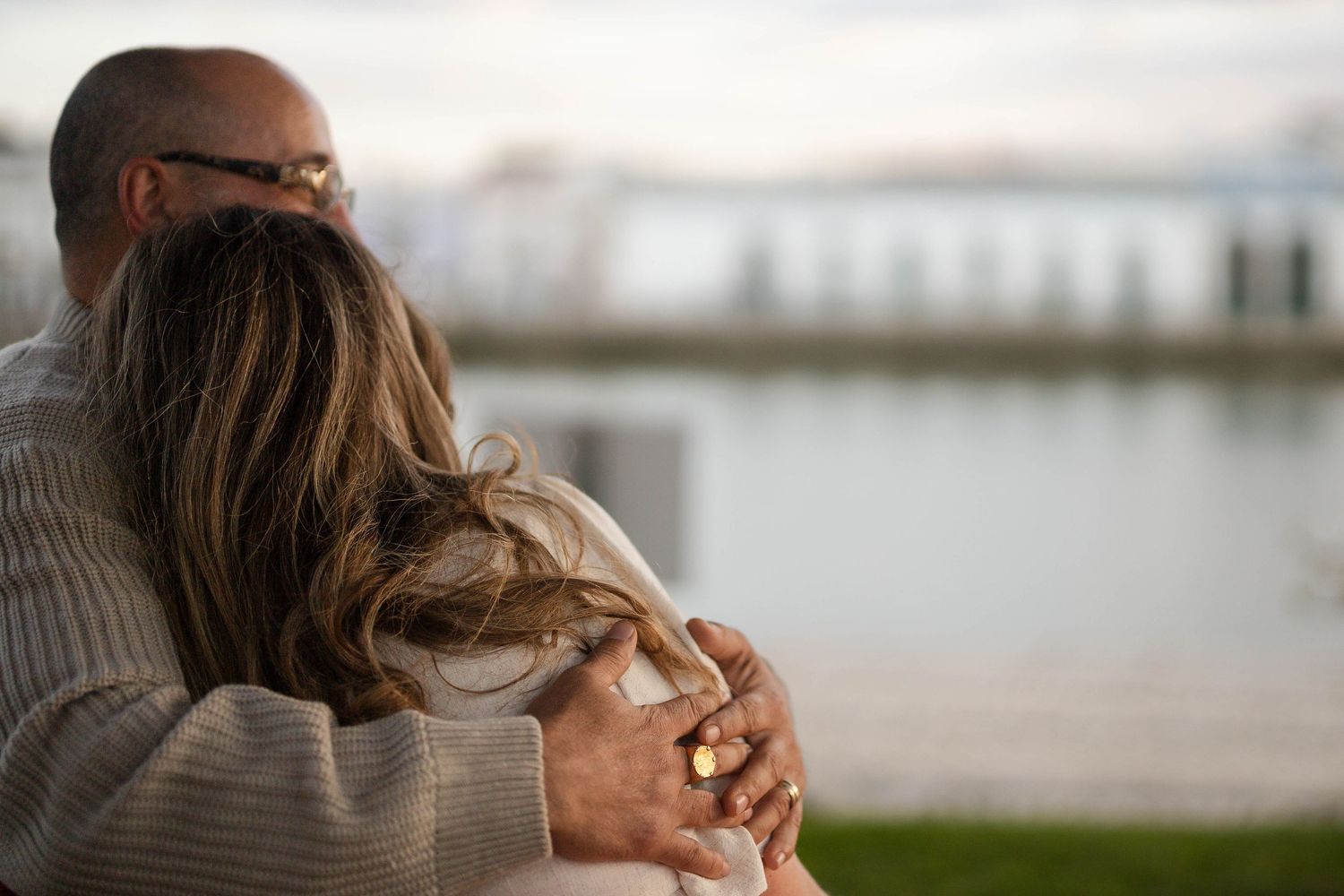 Couple sharing a warm embrace outdoors near a waterfront, viewed from behind on a cloudy day.