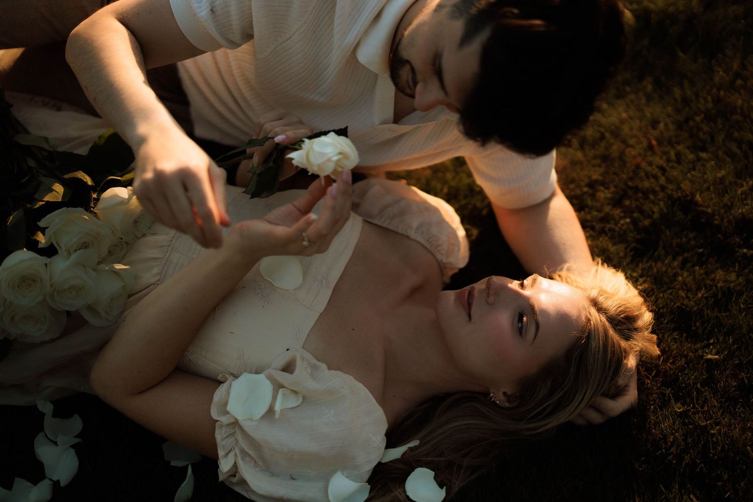 A woman in a white dress lies on grass surrounded by white flowers while a person leans over her tenderly.