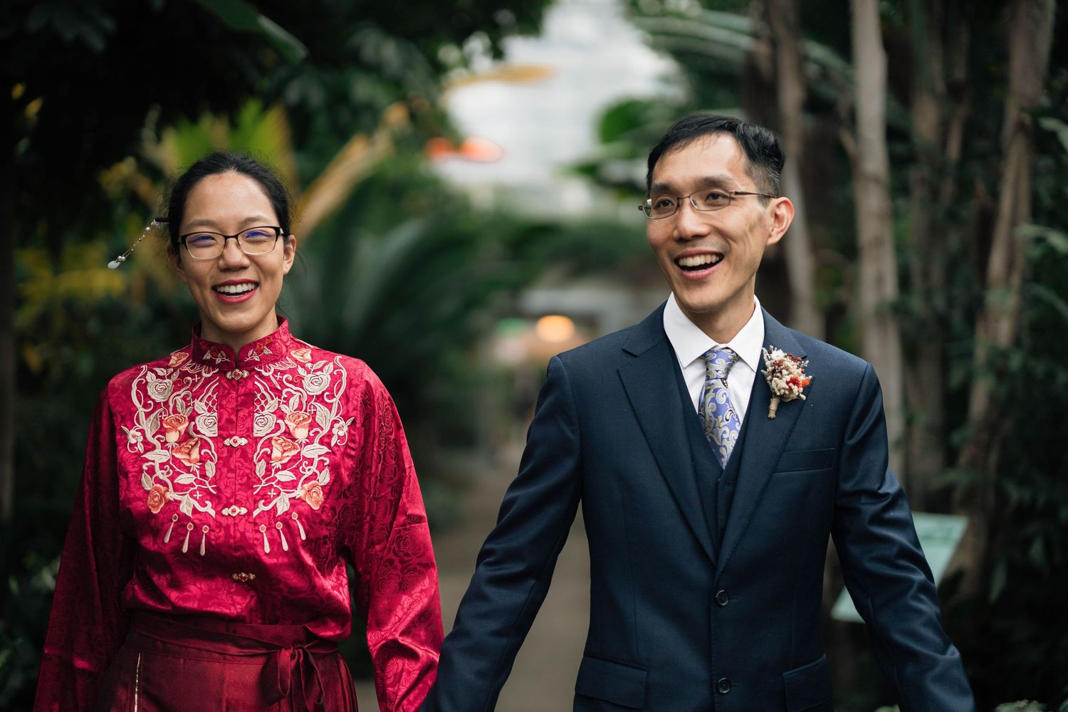 Smiling Asian couple in wedding attire, woman in red embroidered qipao and man in navy suit with boutonniere, outdoors.