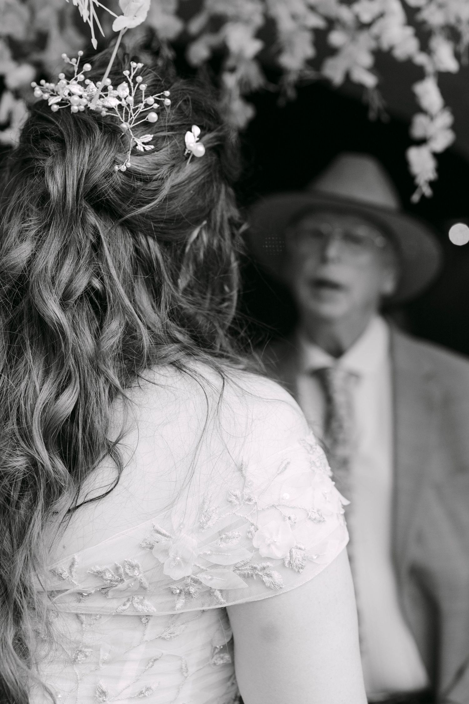 Black and white photo of a bride with floral hair accessories facing an older man in a hat at a wedding ceremony.