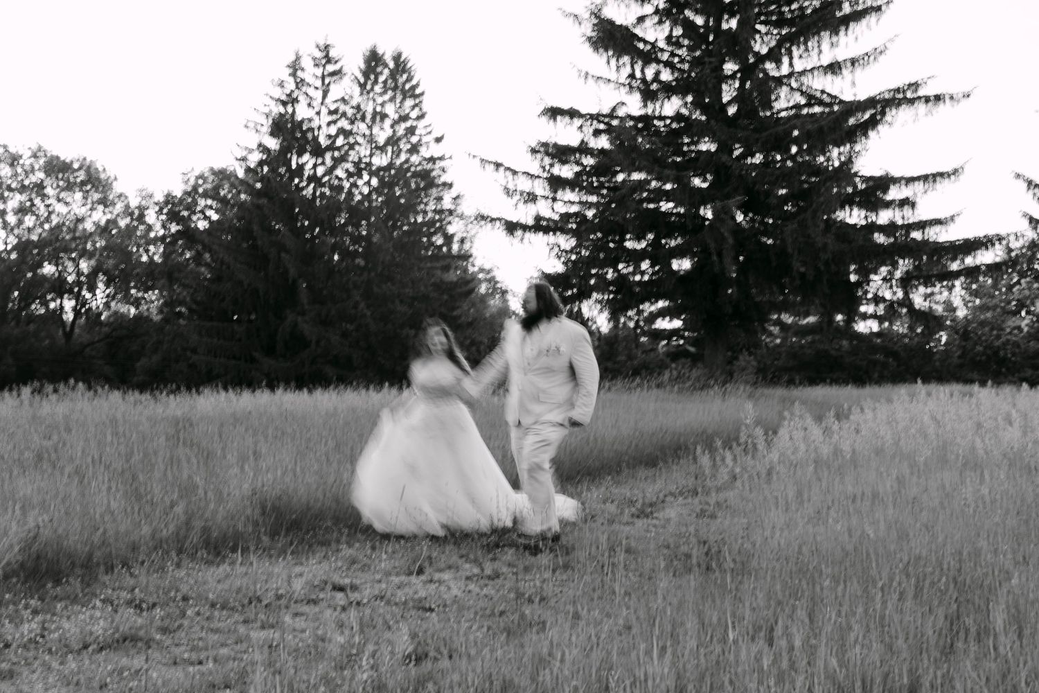 Black and white photo of a bride and groom holding hands, running through a field with tall trees in the background.