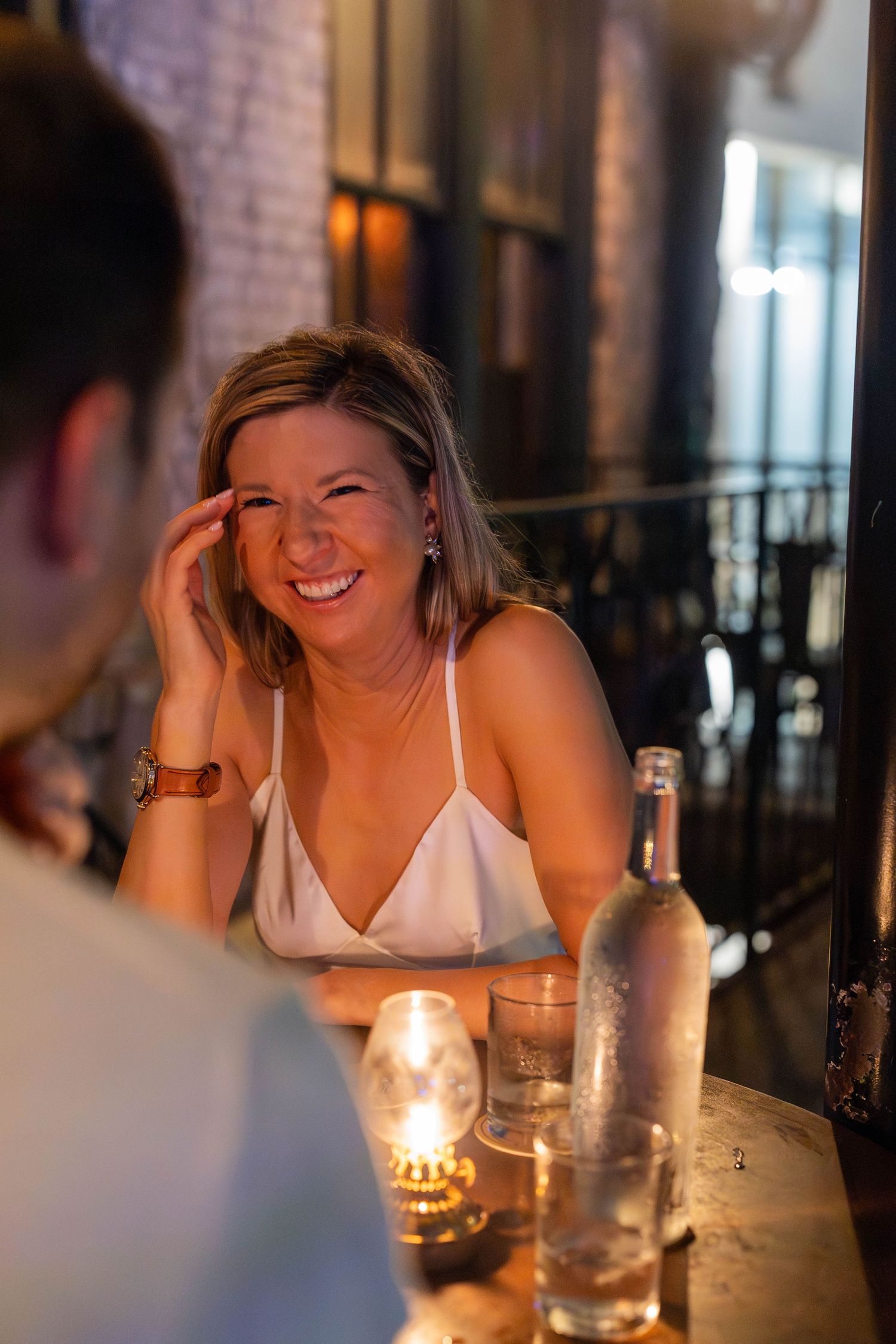 Smiling blonde woman laughing at a candlelit restaurant table with a bottle and glasses nearby.