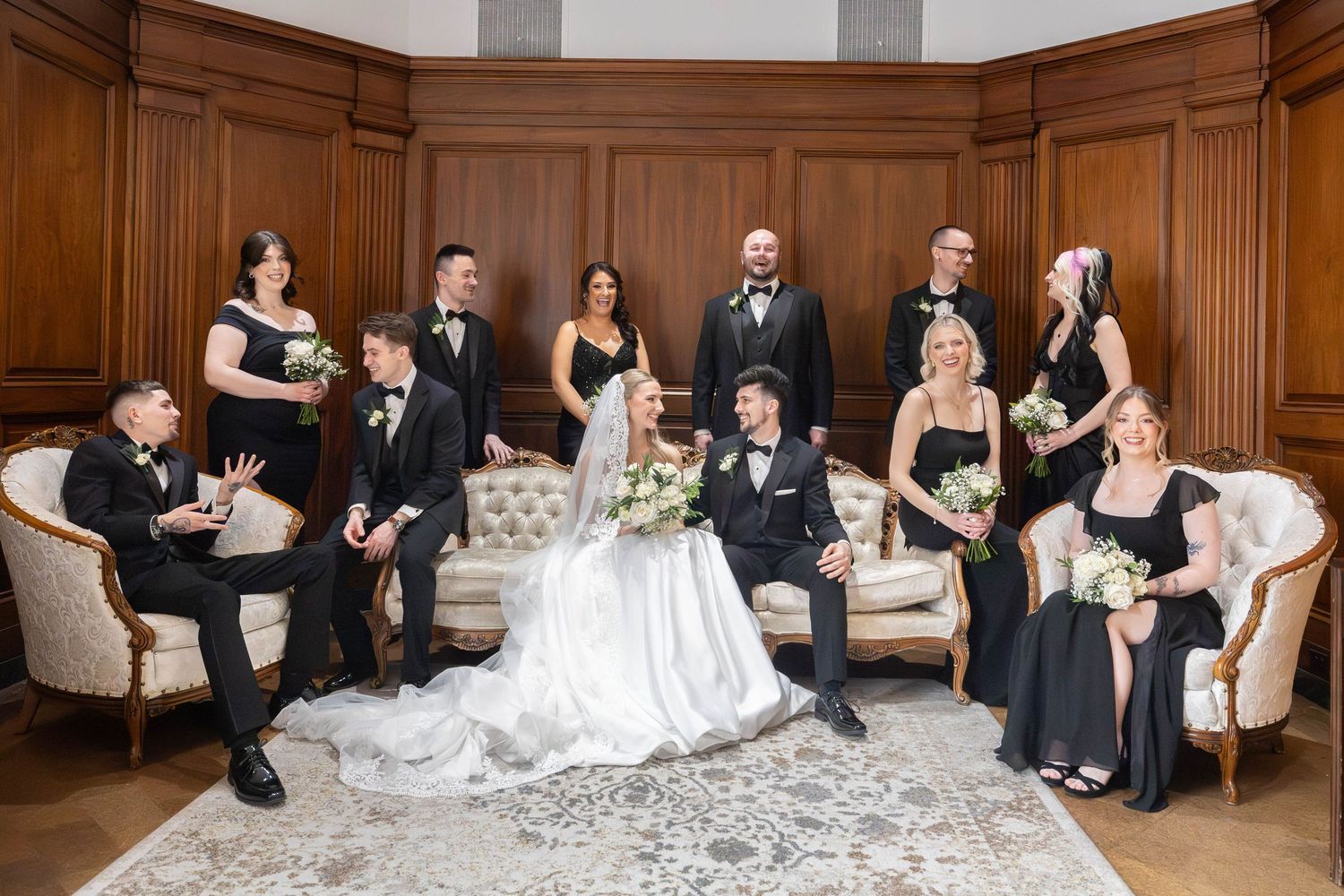 Wedding party in black attire poses with bride and groom on elegant sofas in a wood-paneled room.