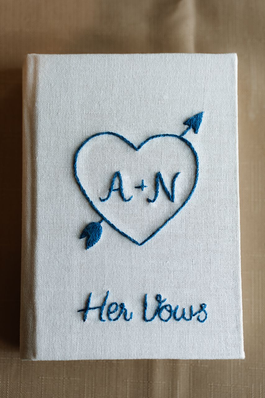 White embroidered vow book with blue heart, arrow, and initials A+N reading Her Vows for a wedding.