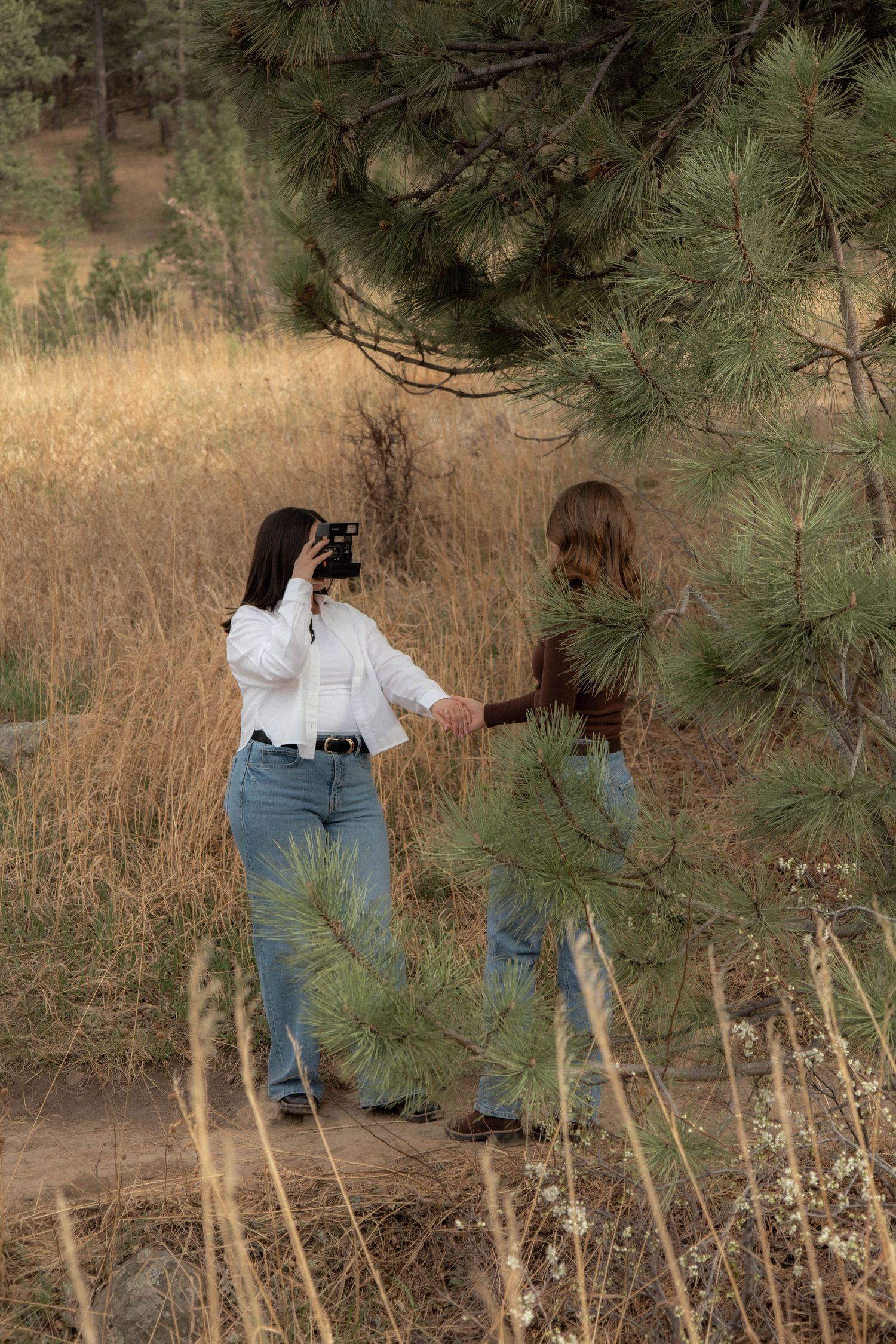a woman taking a photo of another woman holding hands with a green tree branch framing them in Boulder, Colorado