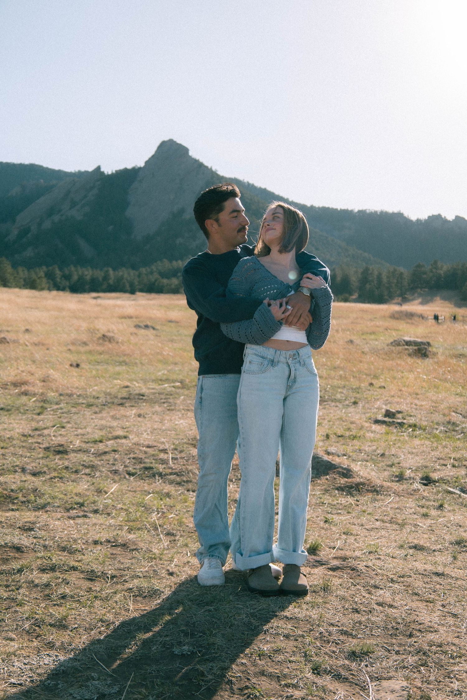 A woman standing in front of a man with his arms wrapped around her in a grassy field in Boulder, Colorado