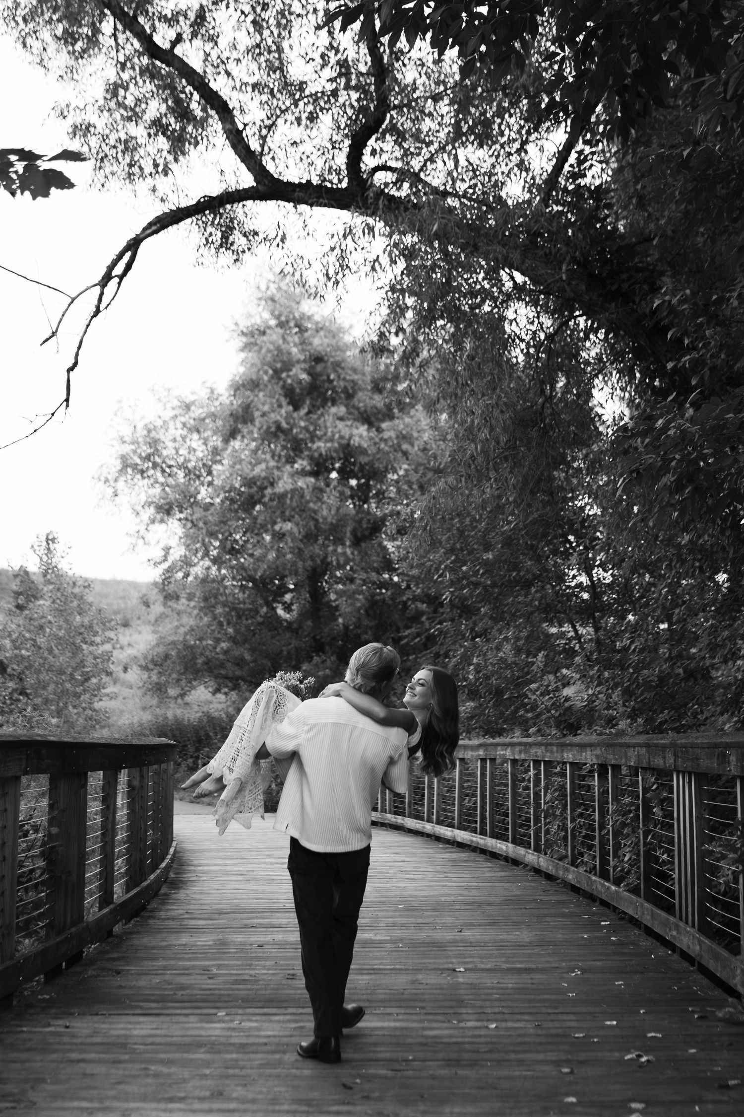Black and white photo of a groom carrying his bride on a wooden bridge surrounded by lush trees.