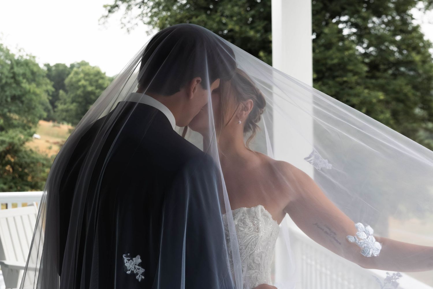 Bride and groom sharing an intimate moment under a flowing bridal veil on an outdoor porch.