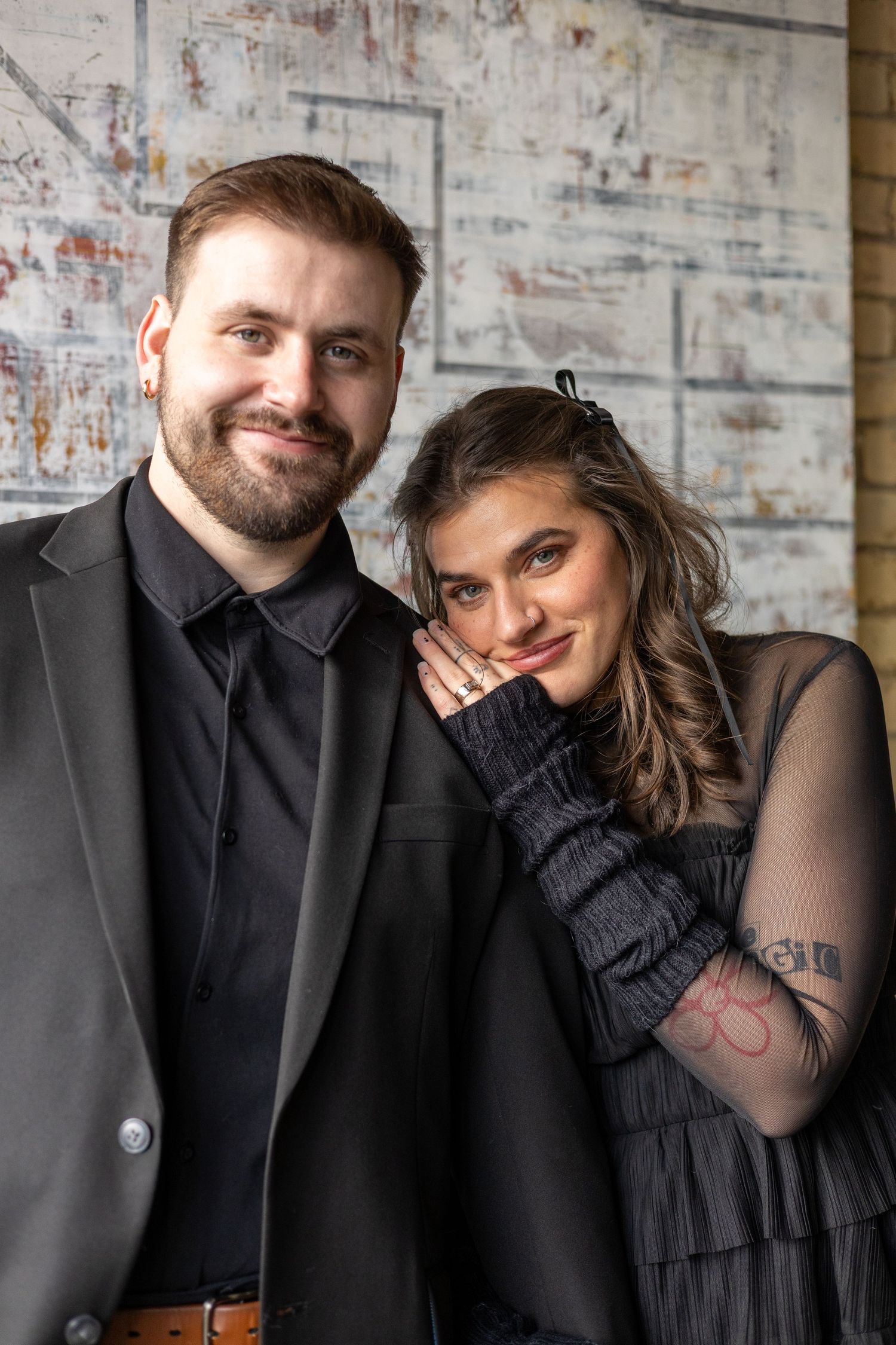 Smiling bearded man in black suit poses with brunette woman in sheer black dress for engagement photo.