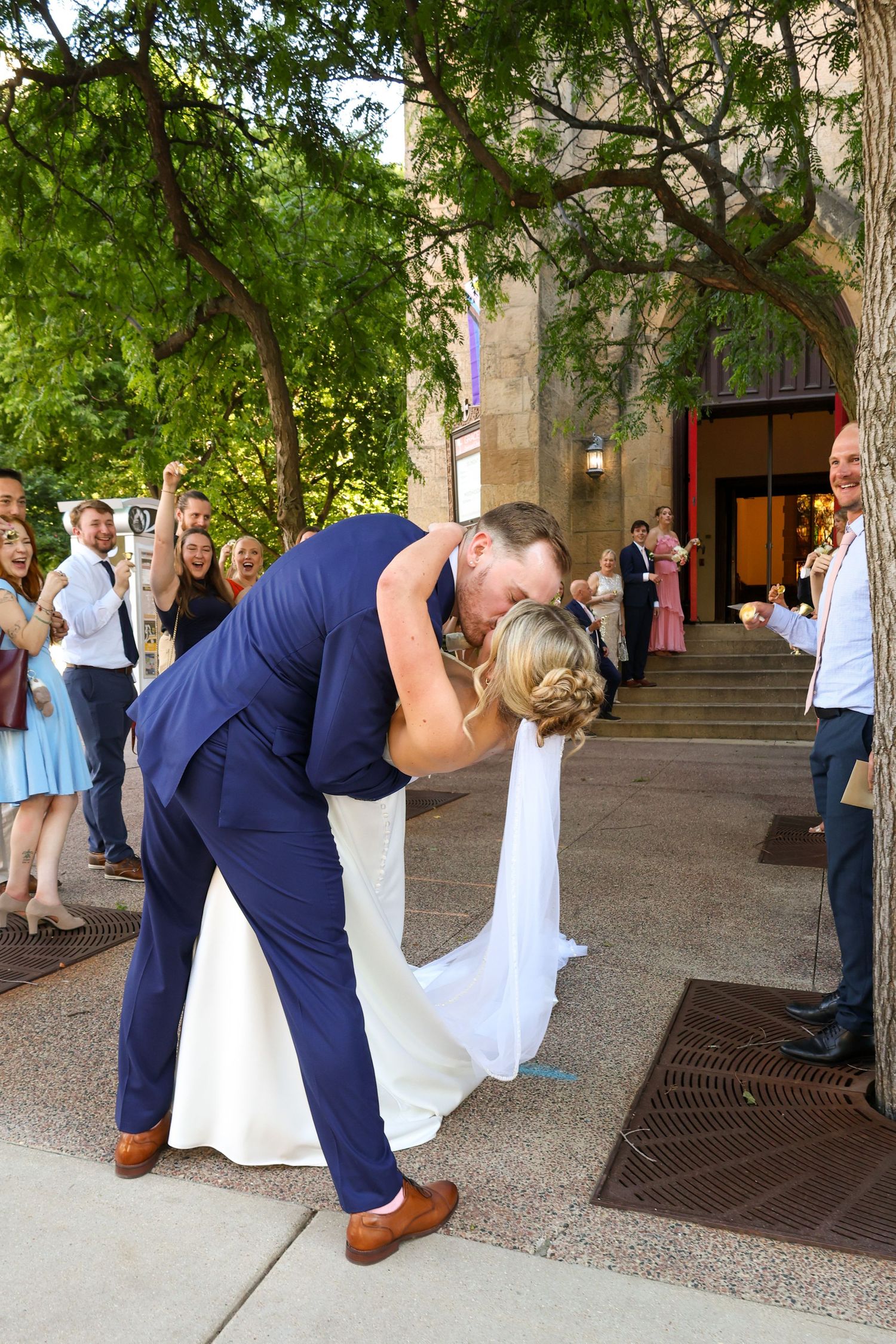 Groom in navy suit dips and kisses bride in white gown outside wedding venue as guests cheer.