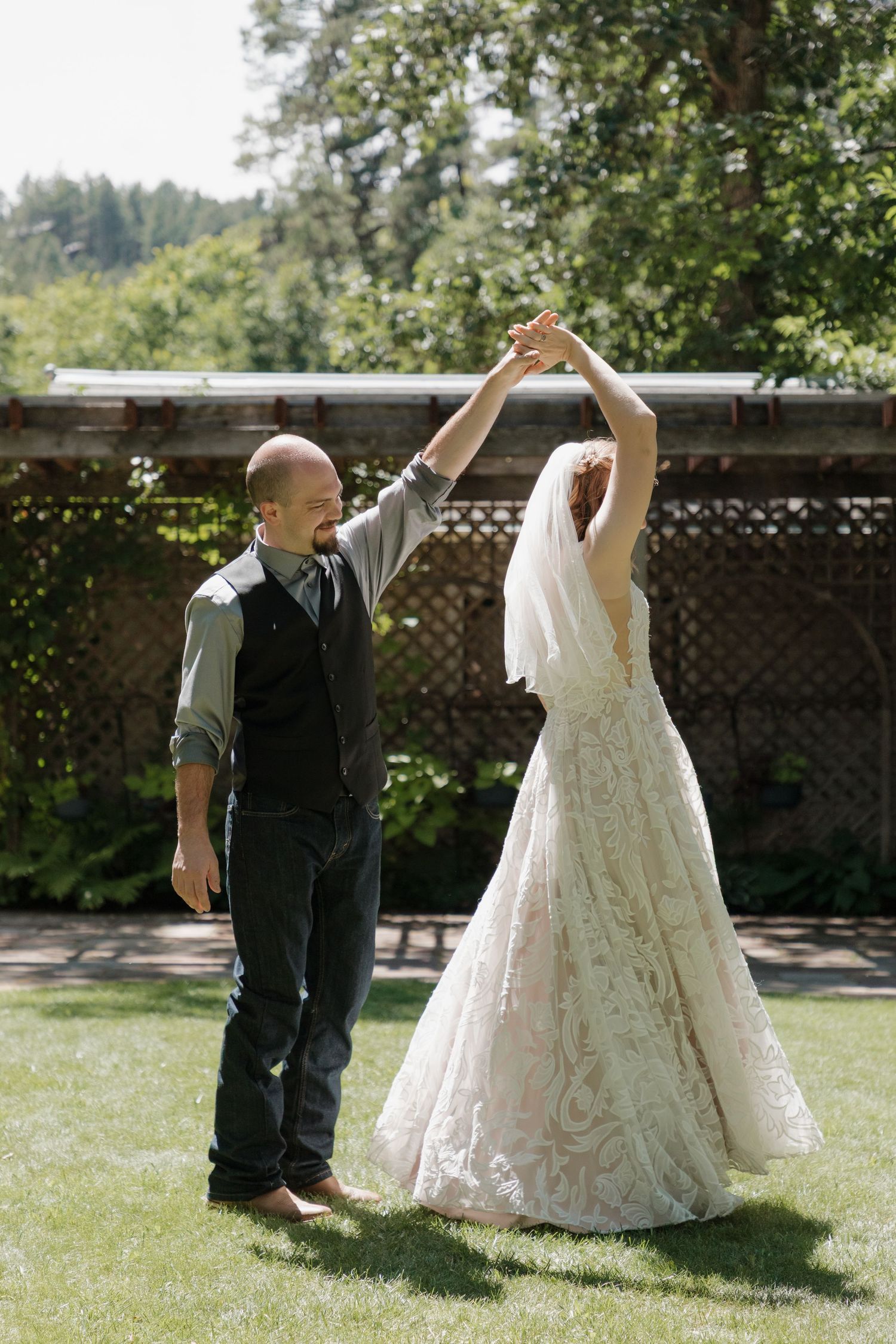 A wedding couple dances together on a sunny outdoor lawn with trees in the background.