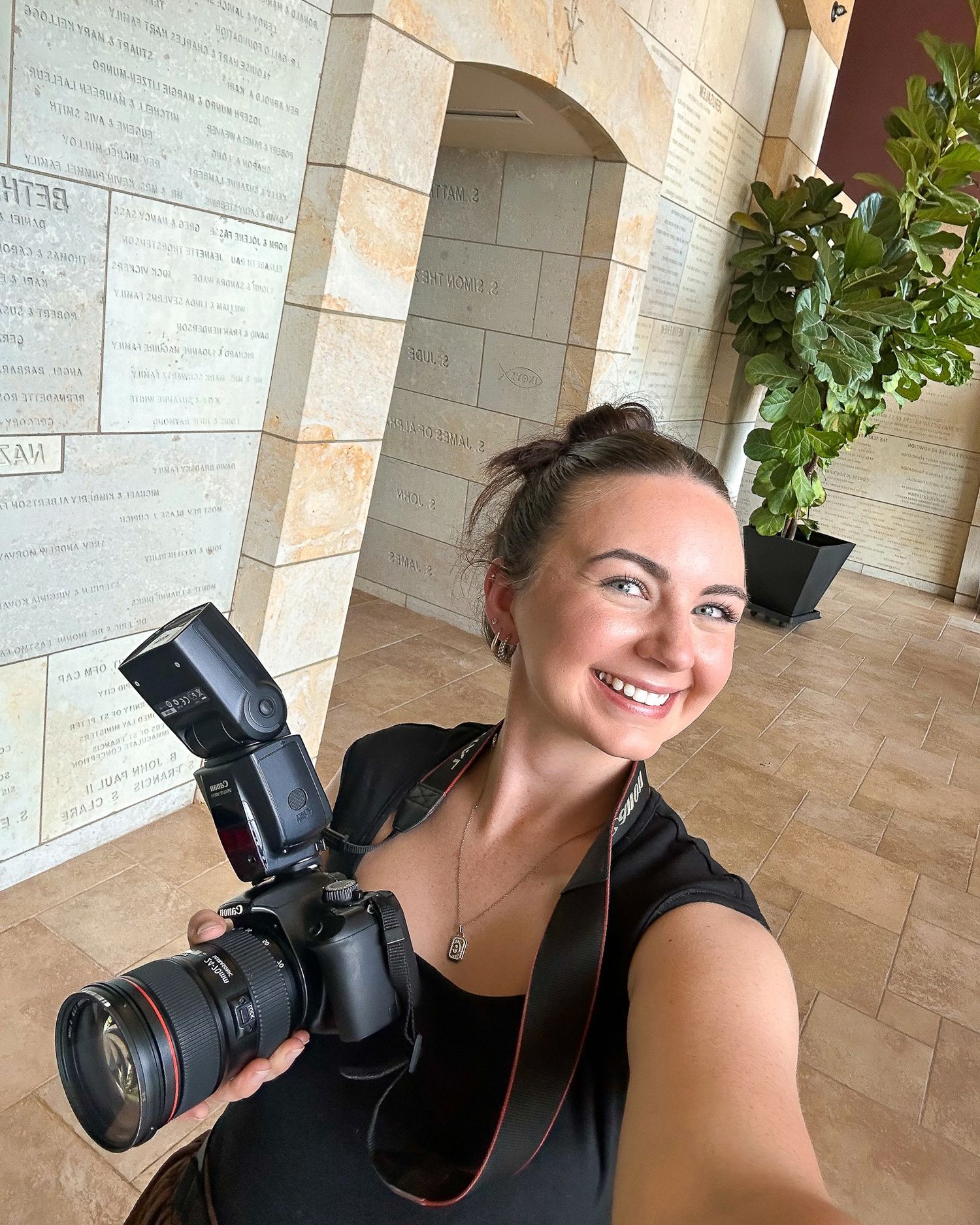 Professional photographer smiling while holding camera equipment in a bright hallway.