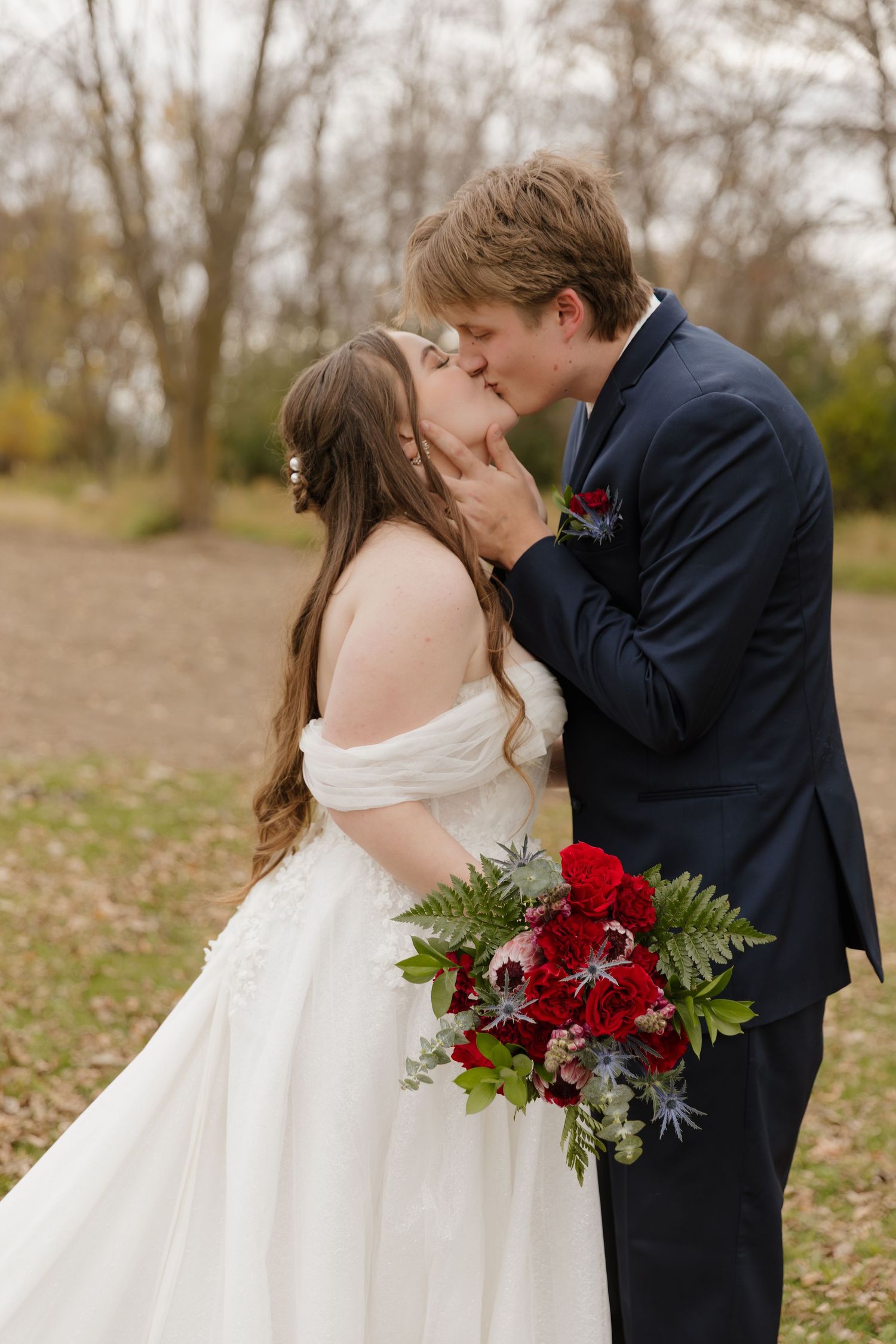 Romantic wedding couple sharing a tender moment with red bouquet in an autumn setting.