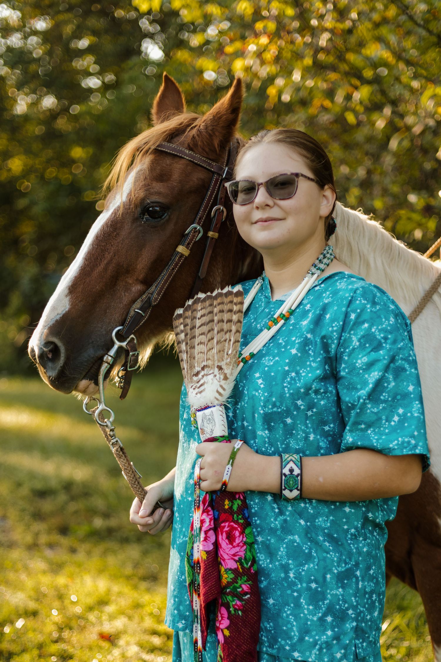 Person in turquoise dress posing with paint horse wearing bridle in autumn setting.
