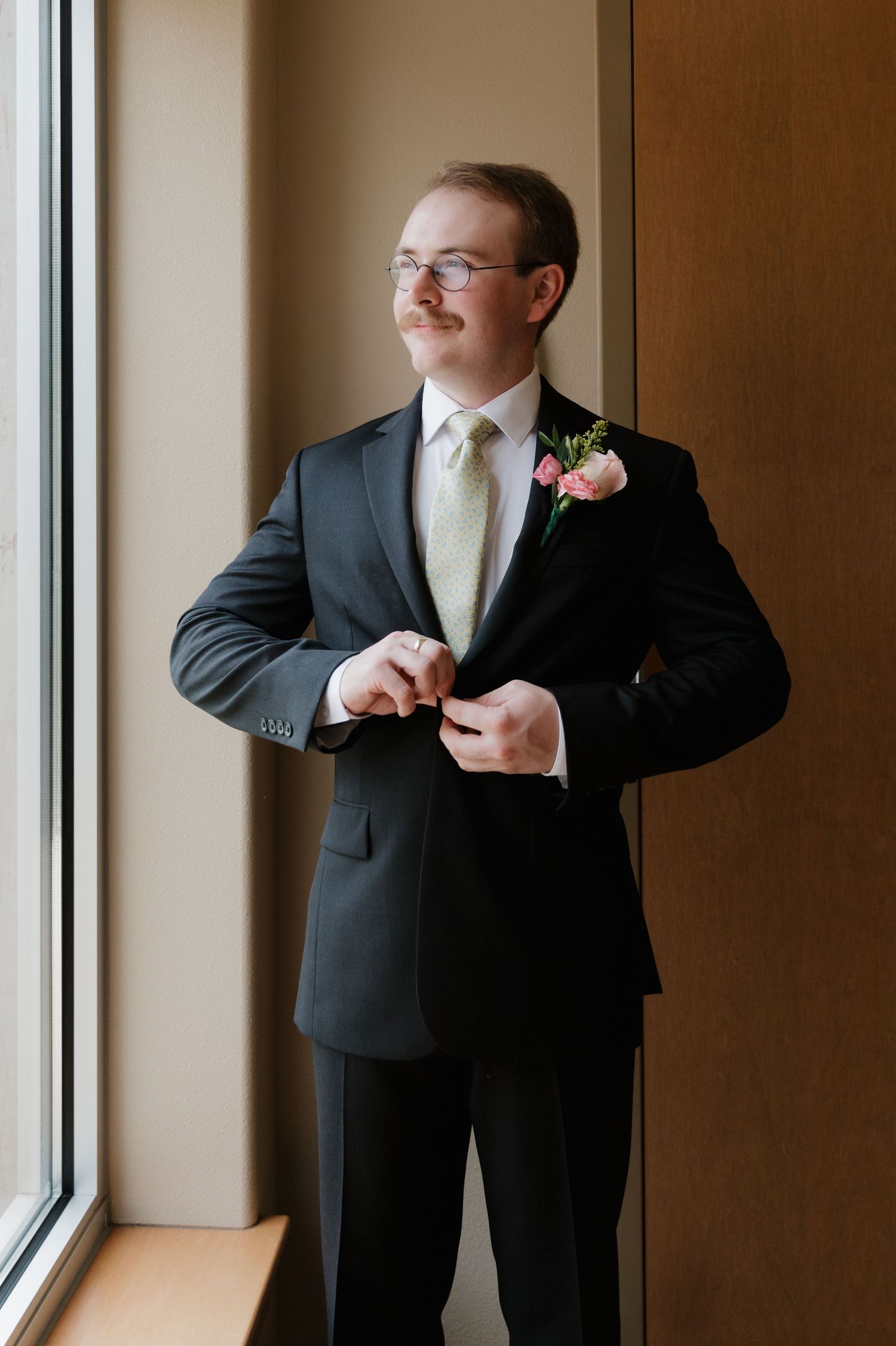 Men in suits adjust their ties while standing near large windows in a formal setting.