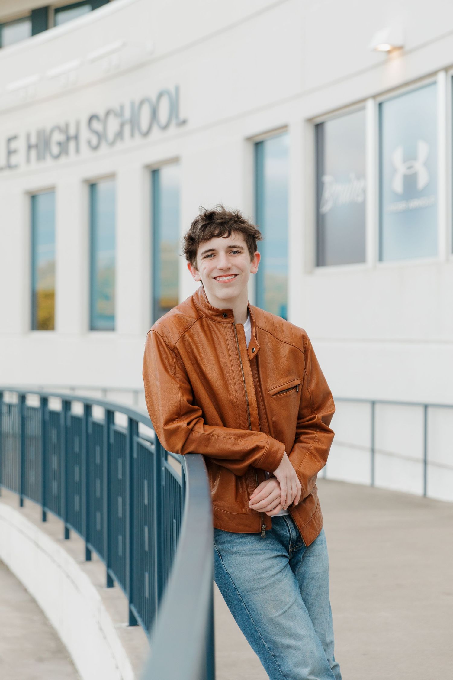 Student in brown jacket and jeans poses casually against white high school building exterior.