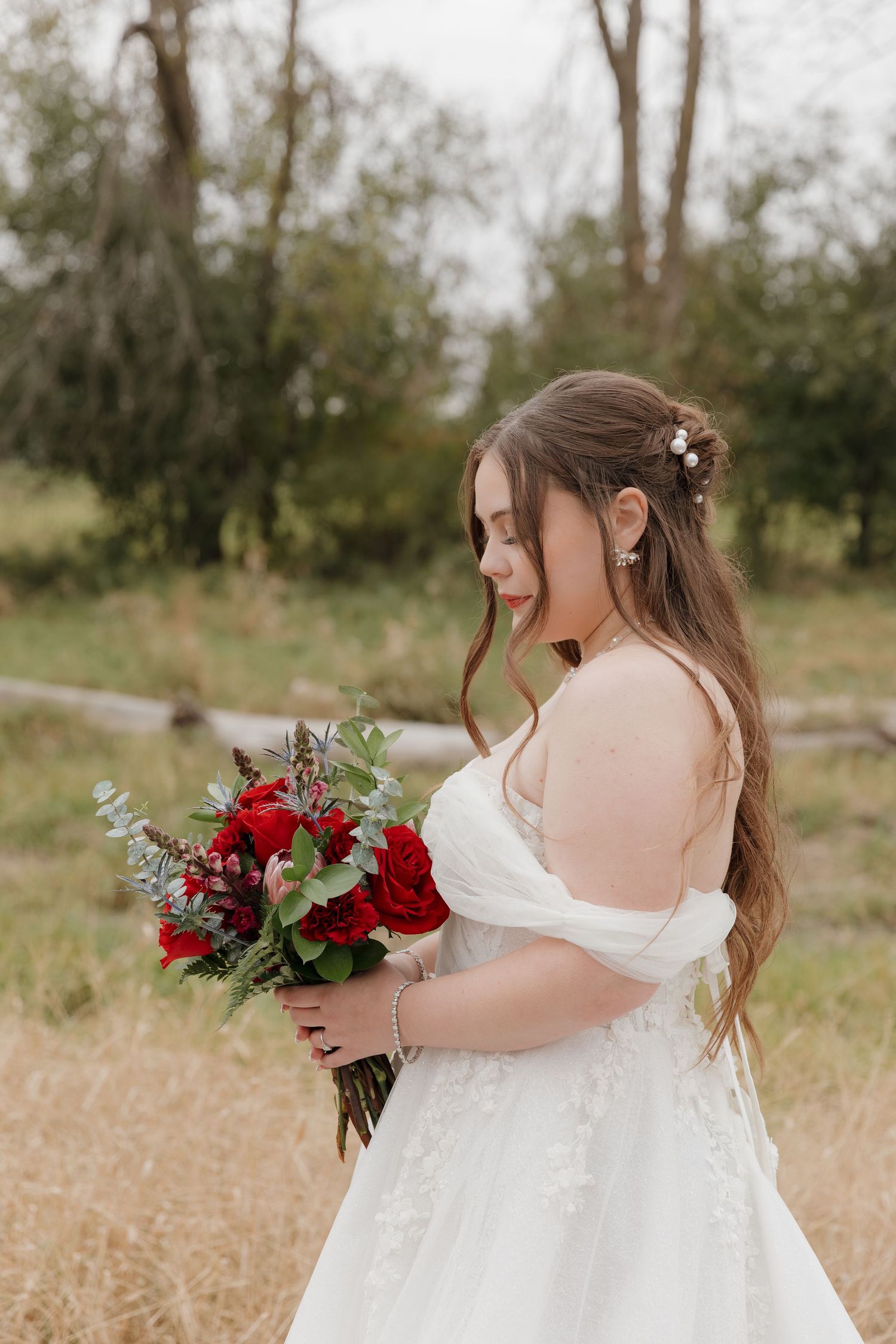 Person in white dress holding red and white floral bouquet in outdoor field.
