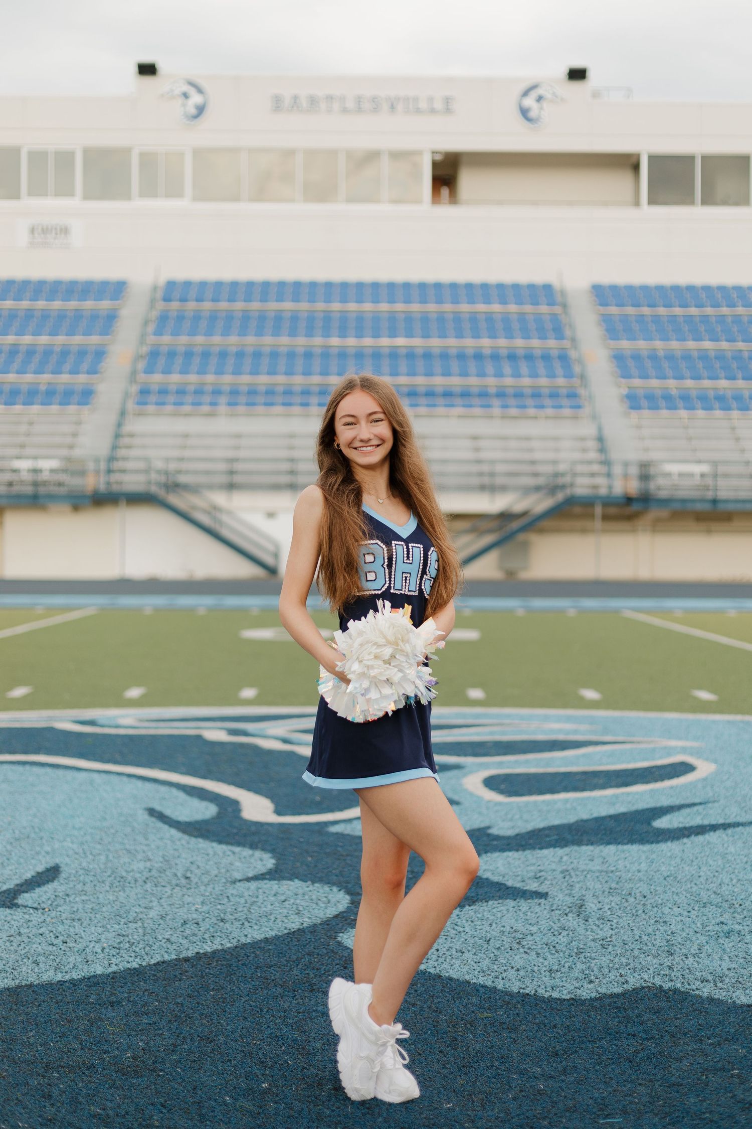 Cheerleader in navy uniform poses with pom poms on football field with stadium seating in background.