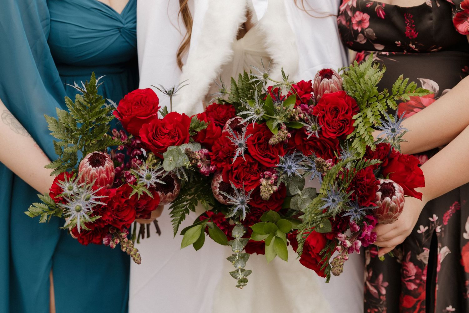 Line of bridesmaids holding lush burgundy and red floral bouquets with greenery.